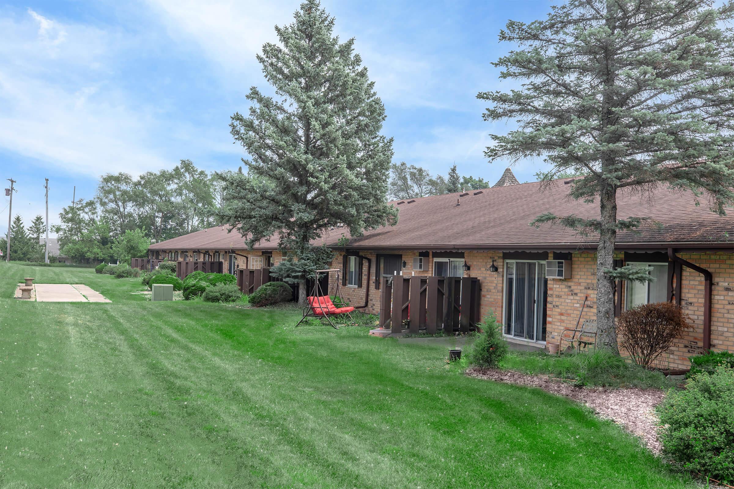 A row of single-story brick townhouses with brown roofs, surrounded by green grass and trees. There are two red lounge chairs on the lawn and a walkway leading towards the townhouses. The sky is partly cloudy, indicating a pleasant day.