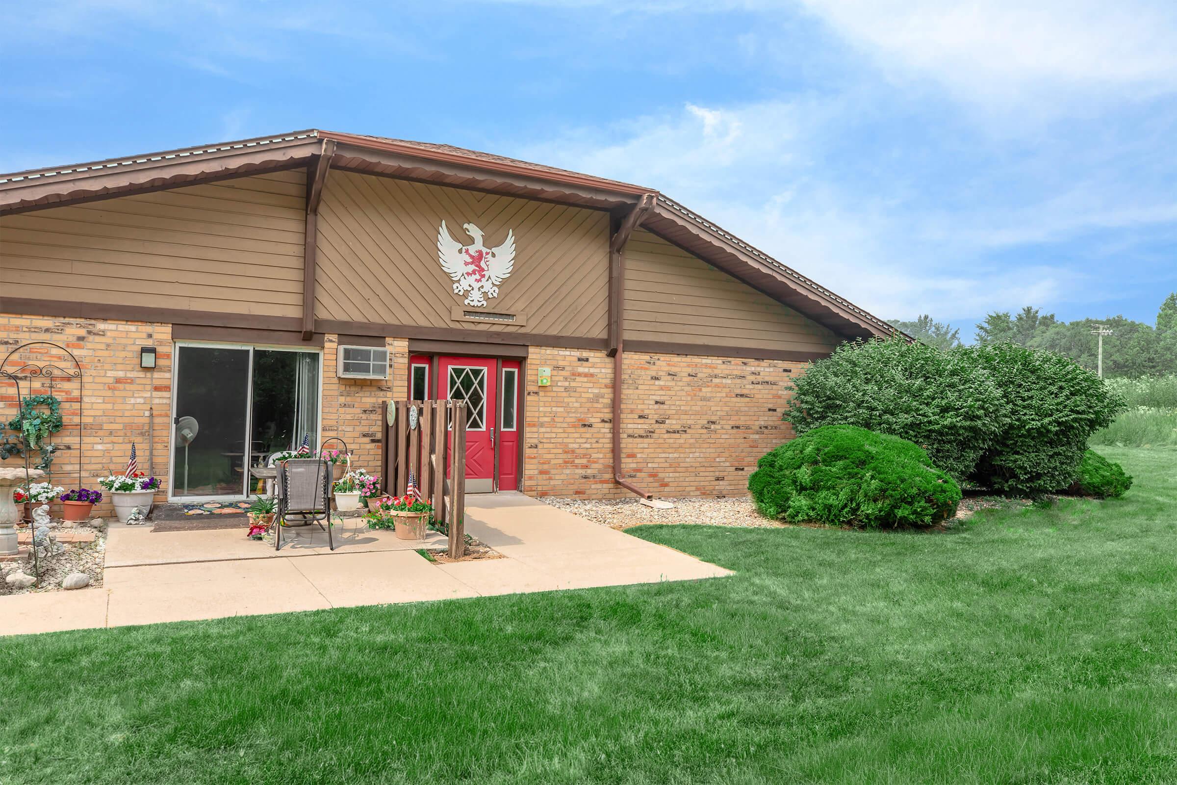 A cozy, single-story home with a rustic design featuring a prominent coat of arms above the entrance. The exterior includes a stone path leading to a red door, surrounded by well-maintained greenery and flower pots. Lush grass and a blue sky create a serene atmosphere.