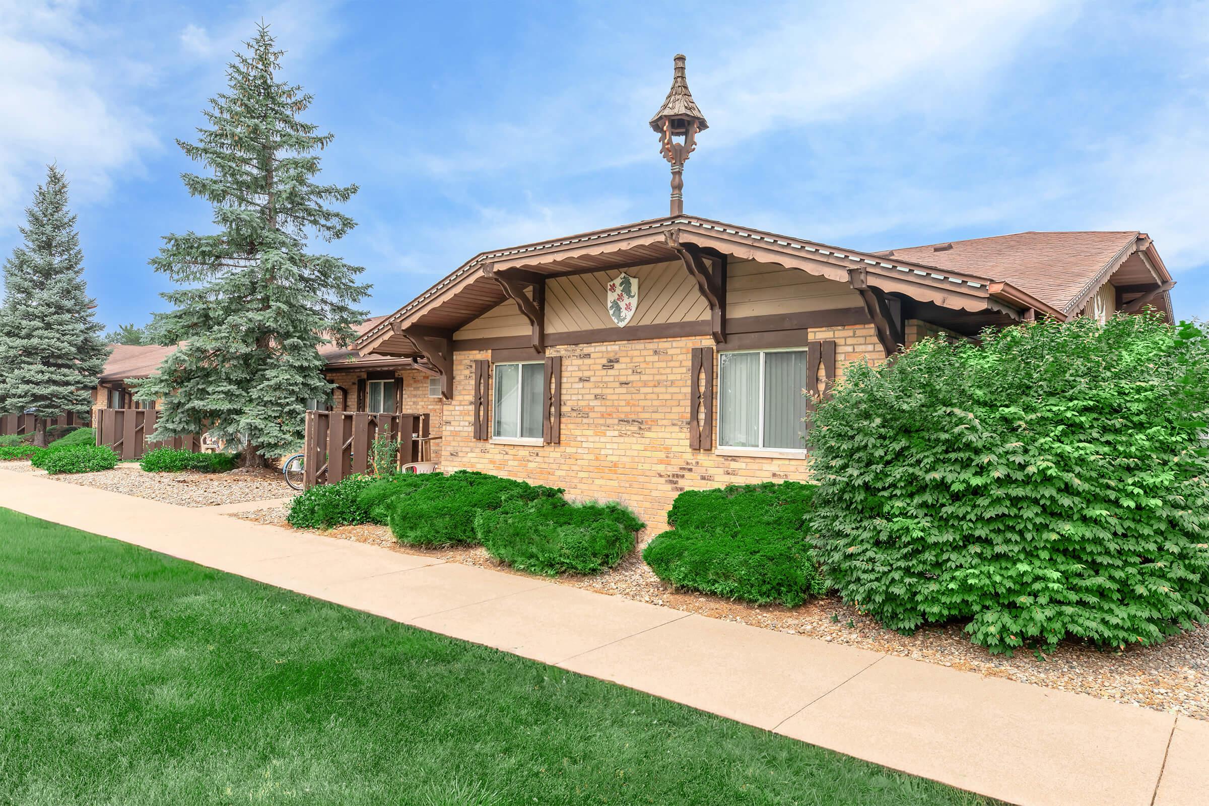 A well-maintained brick building with decorative elements, nestled among green bushes and trees. A paved walkway leads to the entrance, and the sky is blue with scattered clouds, creating a calm and inviting atmosphere.