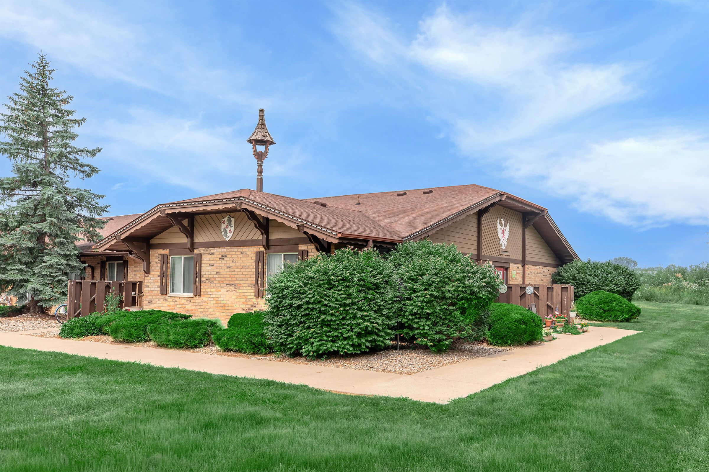 A charming single-story home with a brown and brick exterior, surrounded by well-maintained bushes and grassy lawns. The house features a decorative weathervane on the roof and has a welcoming entrance path lined with greenery against a clear blue sky.