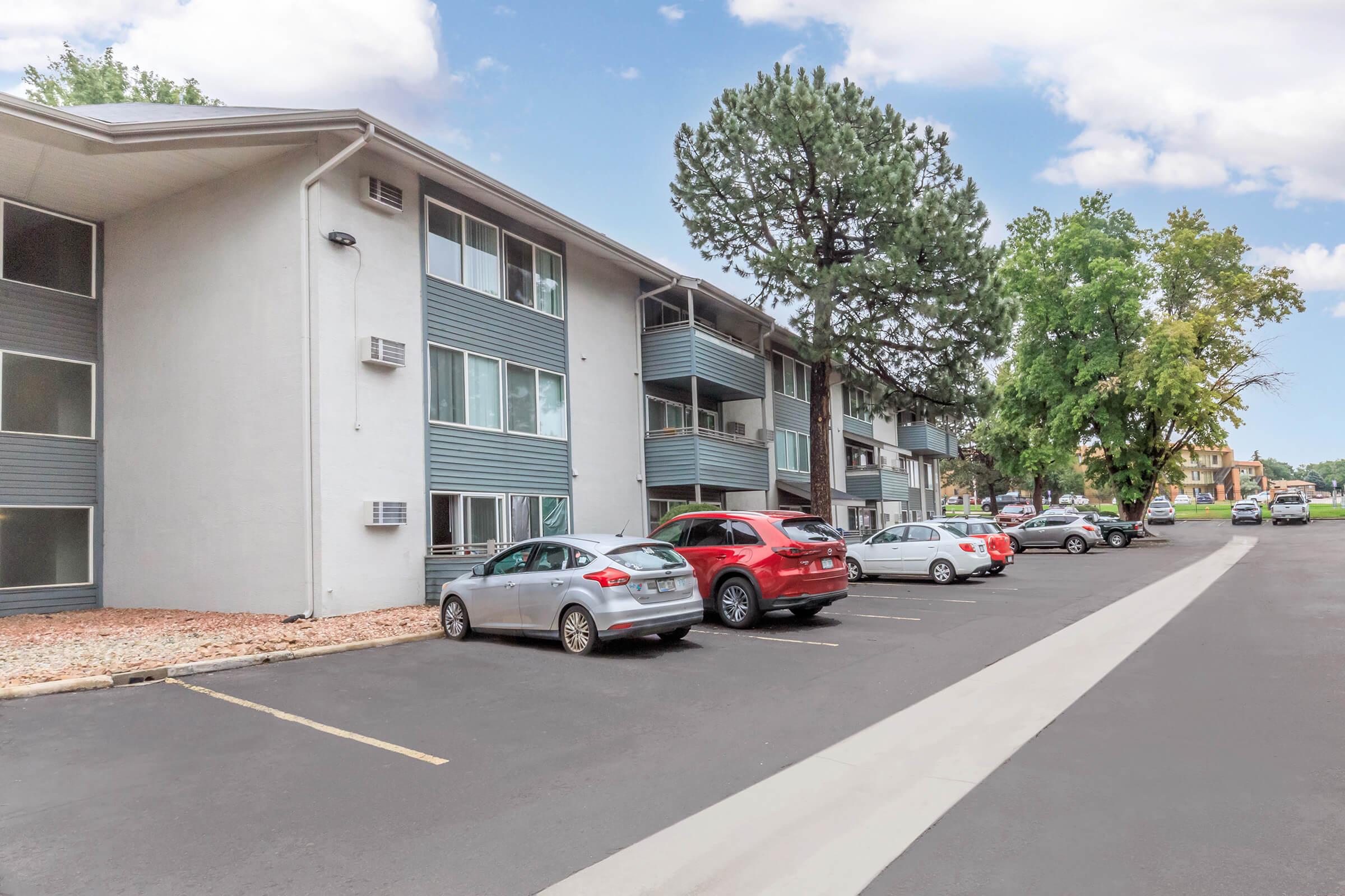 Exterior view of a multi-story apartment building with a parking lot in the foreground. Several cars are parked along a paved driveway, and there are trees nearby. The building features balconies and air conditioning units on its exterior walls, under a clear blue sky.