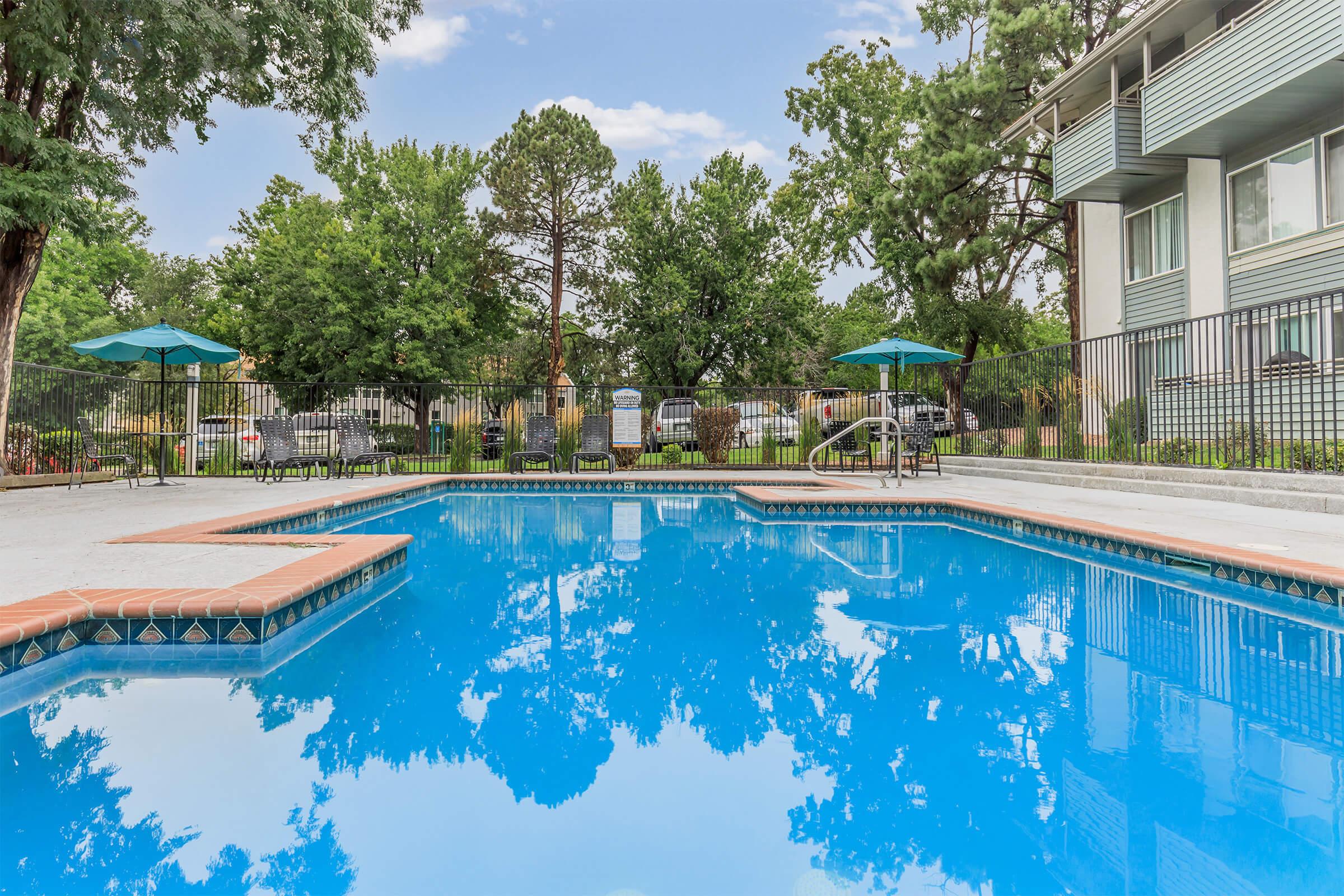 A clear swimming pool surrounded by green trees and umbrellas on a sunny day. The water reflects the sky and surrounding foliage, creating a serene atmosphere. Lounge chairs are arranged near the pool, and there is a fence enclosing the area, adding to the tranquil setting.