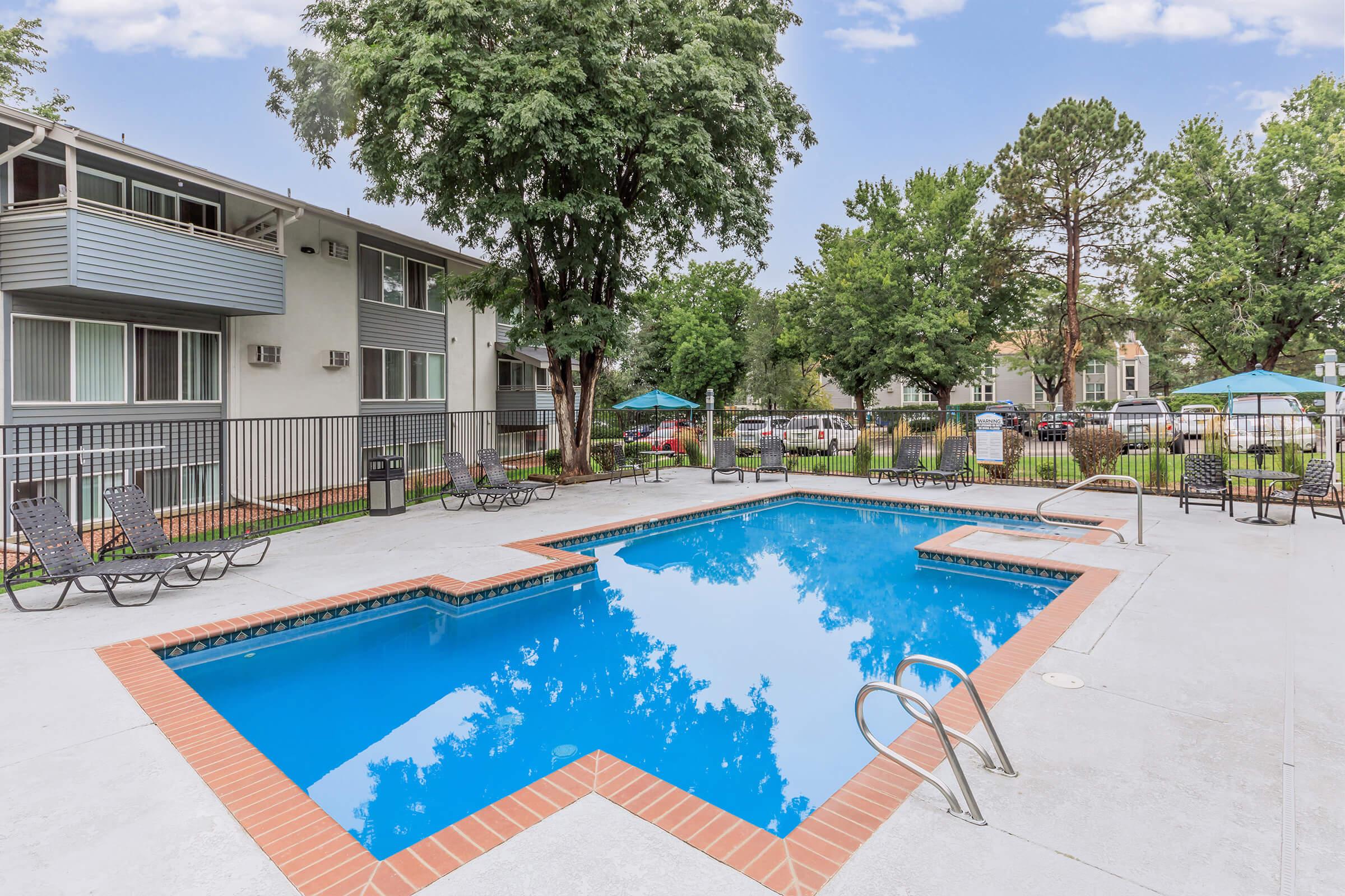 A rectangular swimming pool surrounded by gray concrete, lounge chairs, and umbrellas. Lush trees and greenery are visible in the background, along with apartment buildings. The sky is clear and blue, reflecting in the pool water.