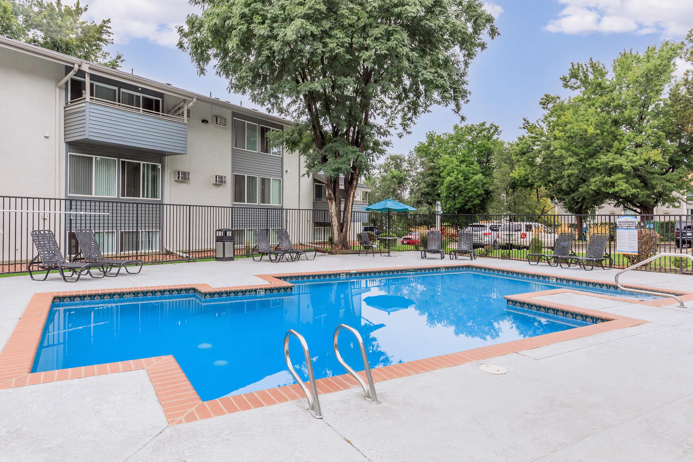 A swimming pool surrounded by lounge chairs and shaded by trees, adjacent to an apartment building. There is an umbrella-covered seating area in the background, and the pool has a clear blue surface reflecting the sky. The area is well-maintained and inviting.