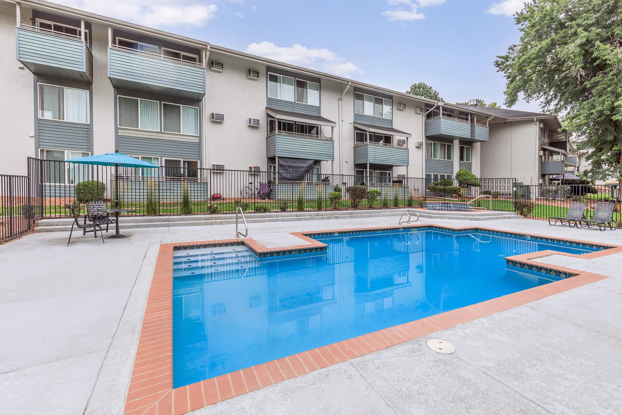 A clear blue swimming pool surrounded by a concrete deck, with lounge chairs and an umbrella nearby. In the background, there's a residential building with multiple balconies and air conditioning units. The area is landscaped with shrubs and small trees, creating a pleasant outdoor atmosphere.