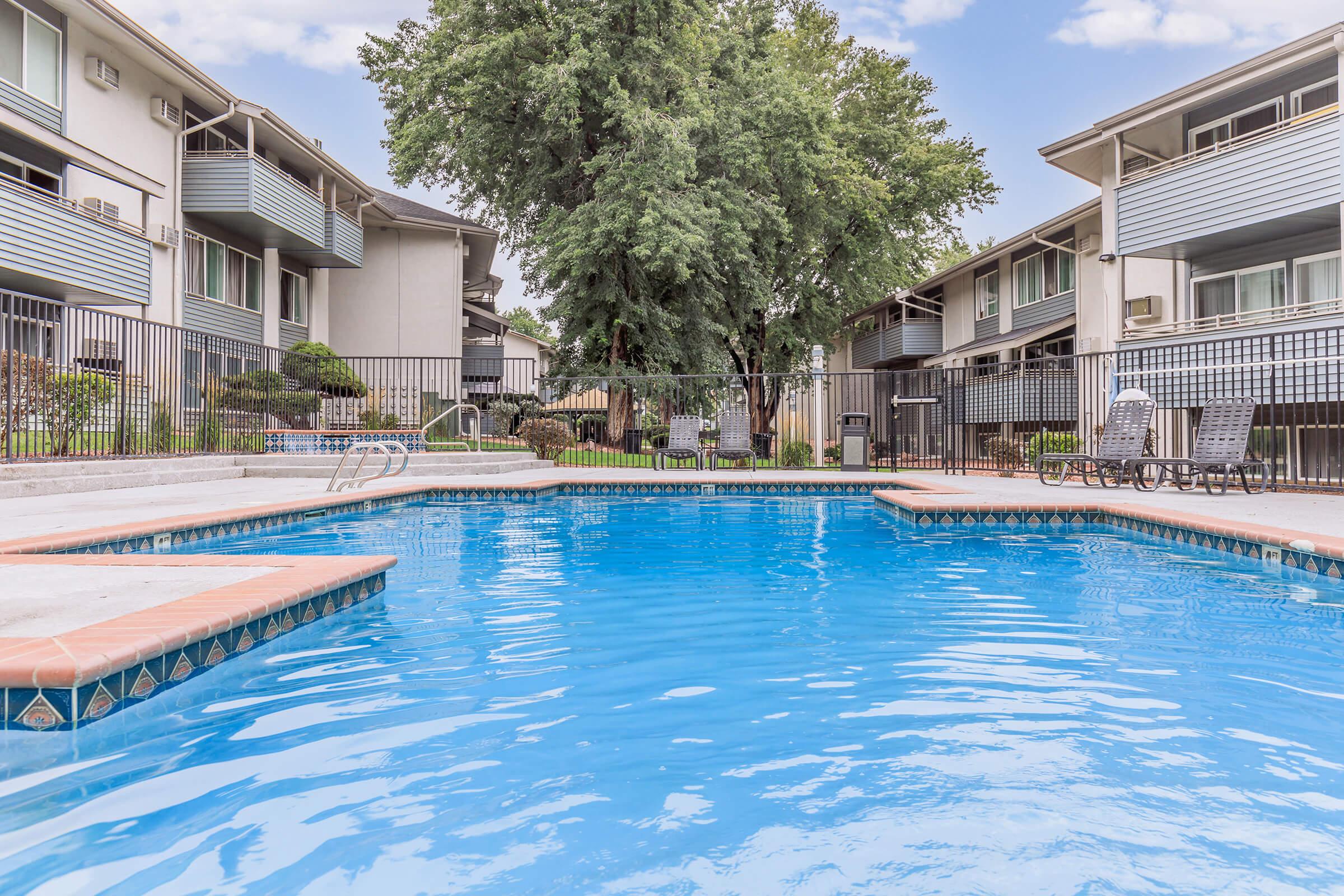 A bright, inviting swimming pool surrounded by lounge chairs and a fence, set against a backdrop of well-maintained apartment buildings and leafy trees. The clear blue water reflects the sky, creating a serene atmosphere perfect for relaxation.