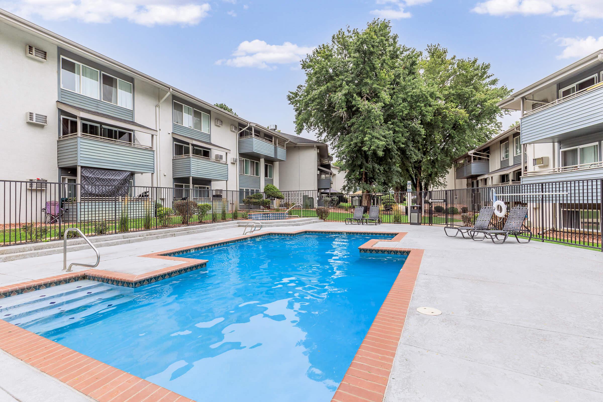 A clear blue swimming pool surrounded by a fenced area, with lounge chairs nearby. In the background, several multi-story apartment buildings are visible, along with green trees and landscaped grass areas. The sky is partly cloudy, creating a pleasant outdoor atmosphere.