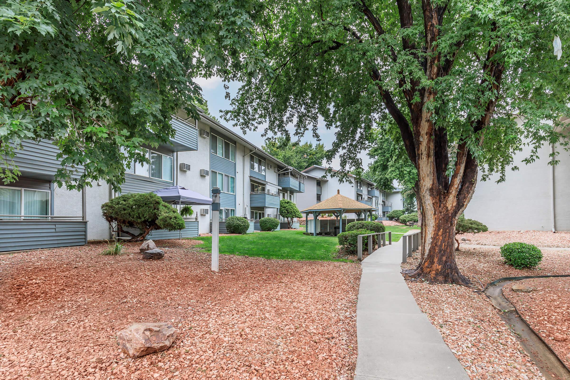 A landscaped area featuring a pathway, a gazebo, and lush green trees surrounding an apartment building complex. The yard is adorned with decorative rocks and manicured shrubs, creating a serene outdoor environment.