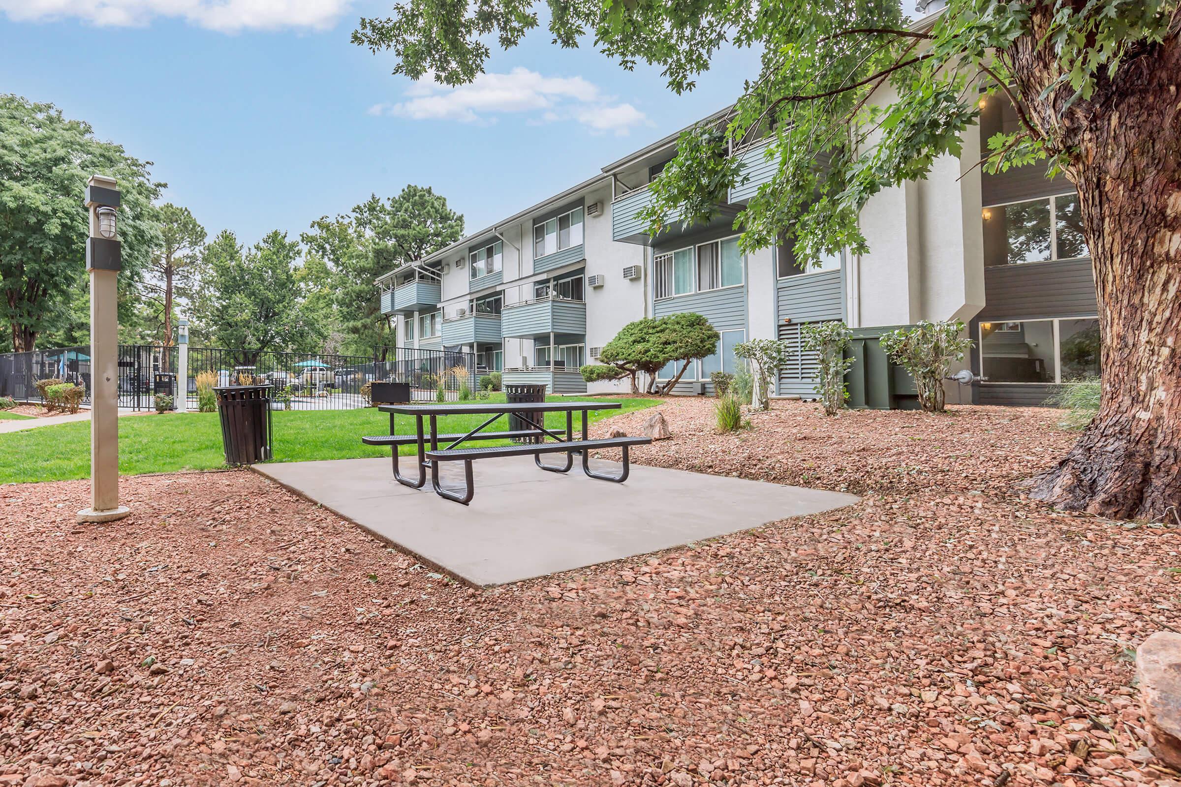 A well-maintained outdoor area featuring a picnic table on a concrete pad surrounded by landscaped gravel and grass. In the background, a multi-story apartment building and trees are visible, creating a peaceful community environment.