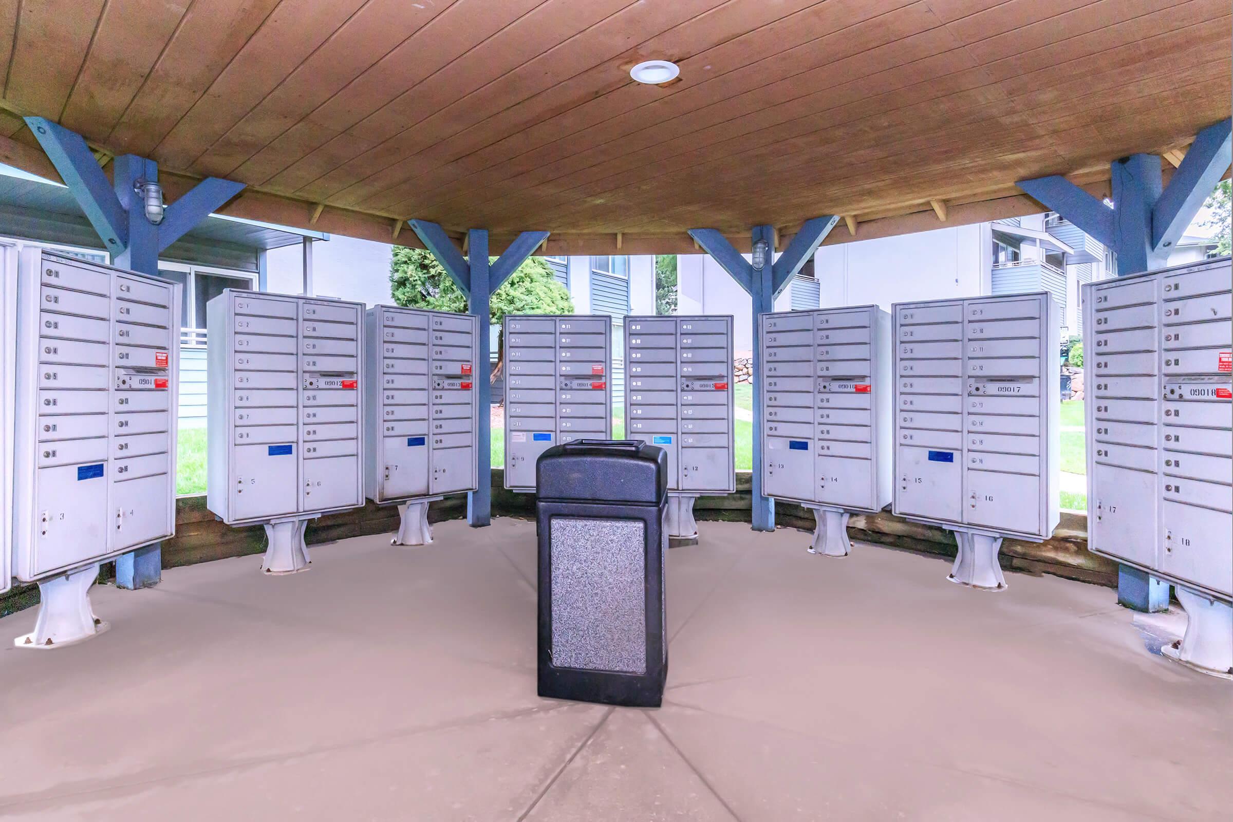 A circular mail kiosk featuring several rows of metal mailboxes, arranged around a central trash bin. The mailboxes have various slot numbers and are covered by a wooden roof, providing shelter. The surrounding area is grassy and features residential buildings in the background.