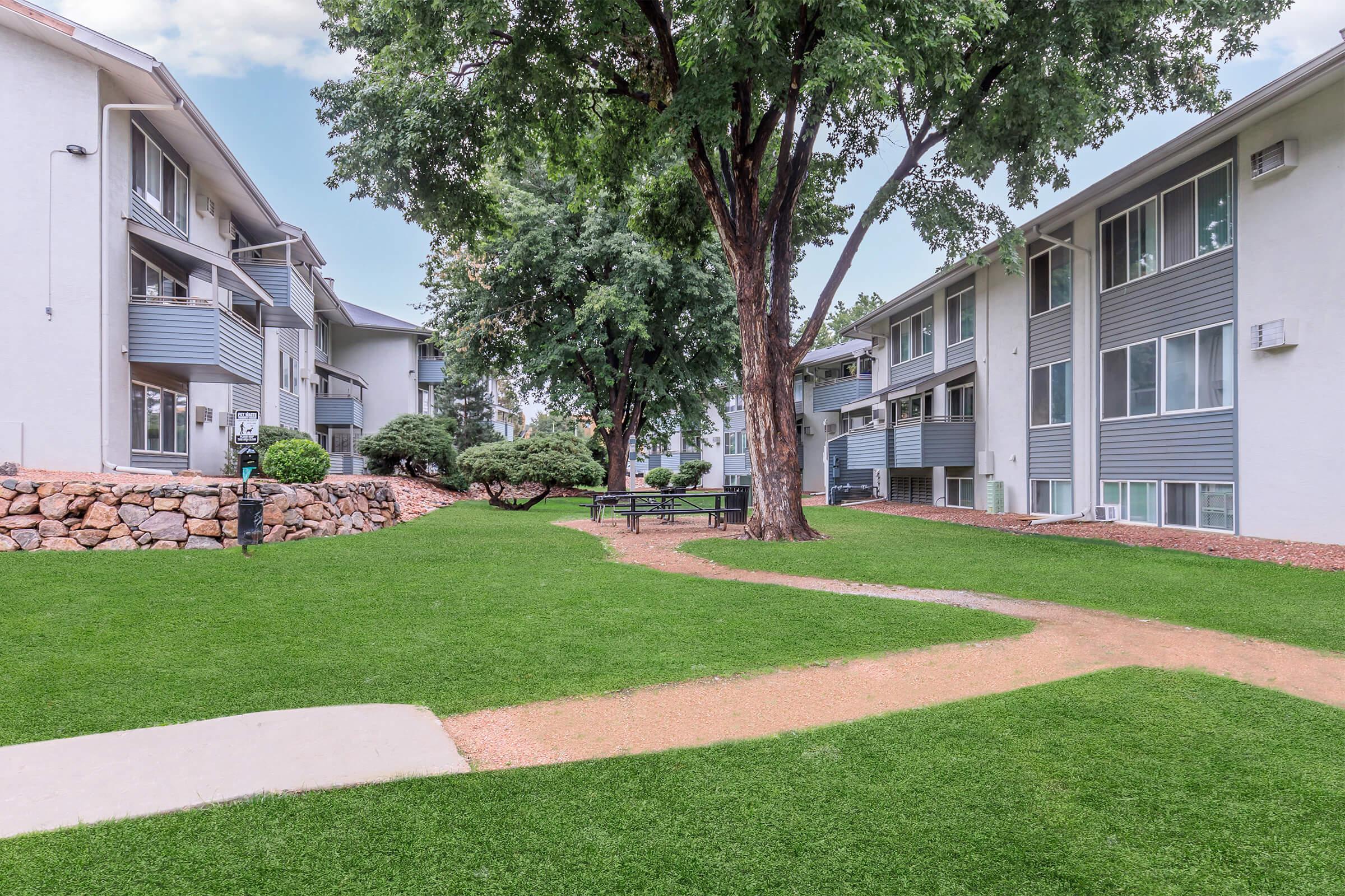 A well-maintained courtyard featuring a grassy area with walking paths, surrounded by two apartment buildings. A large tree provides shade, and there are shrubs and a picnic table visible, creating a welcoming outdoor space for residents.