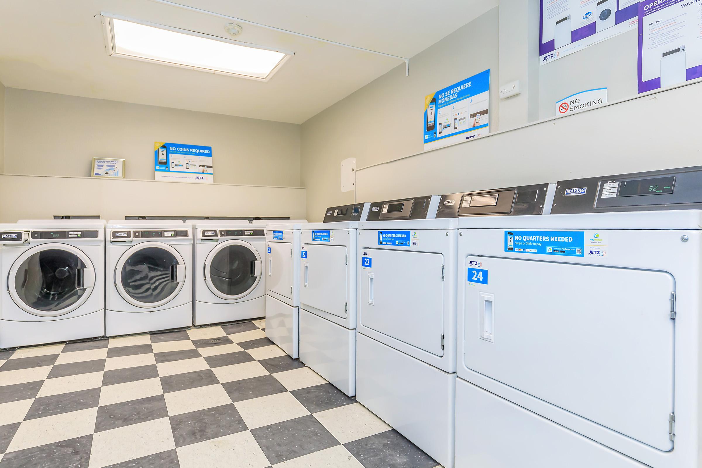 A bright and clean laundromat featuring several white washing machines and dryers lined up against a wall. The floor has a black and white checkerboard pattern. Information posters are visible on the walls, and there's a skylight providing natural light to the space.