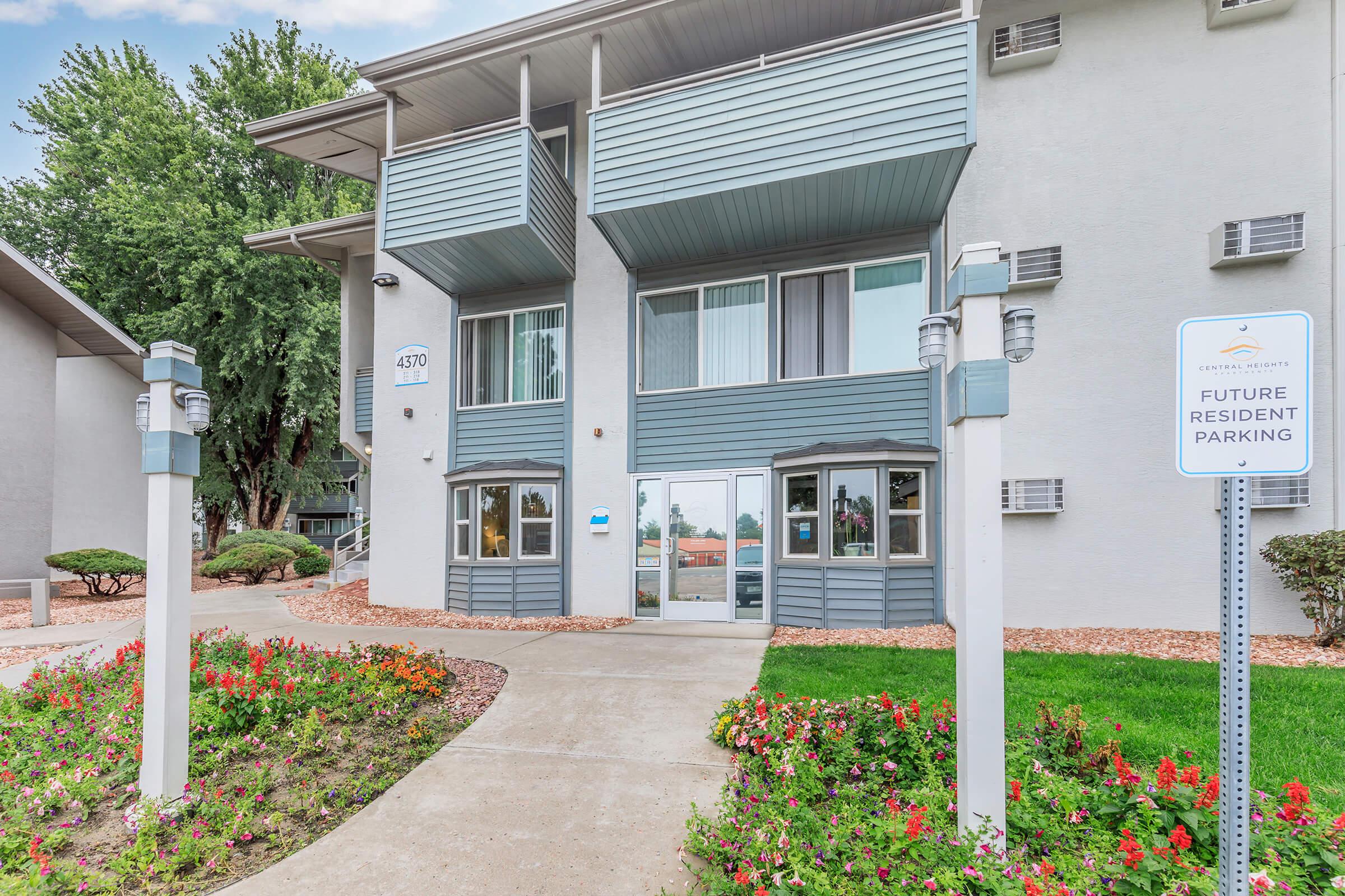 A modern apartment building with a light gray exterior, featuring balconies and large windows. The entrance is flanked by neatly landscaped flower beds, with colorful flowers. A sign indicates "Future Resident Parking." Lush trees are visible in the background under a partly cloudy sky.