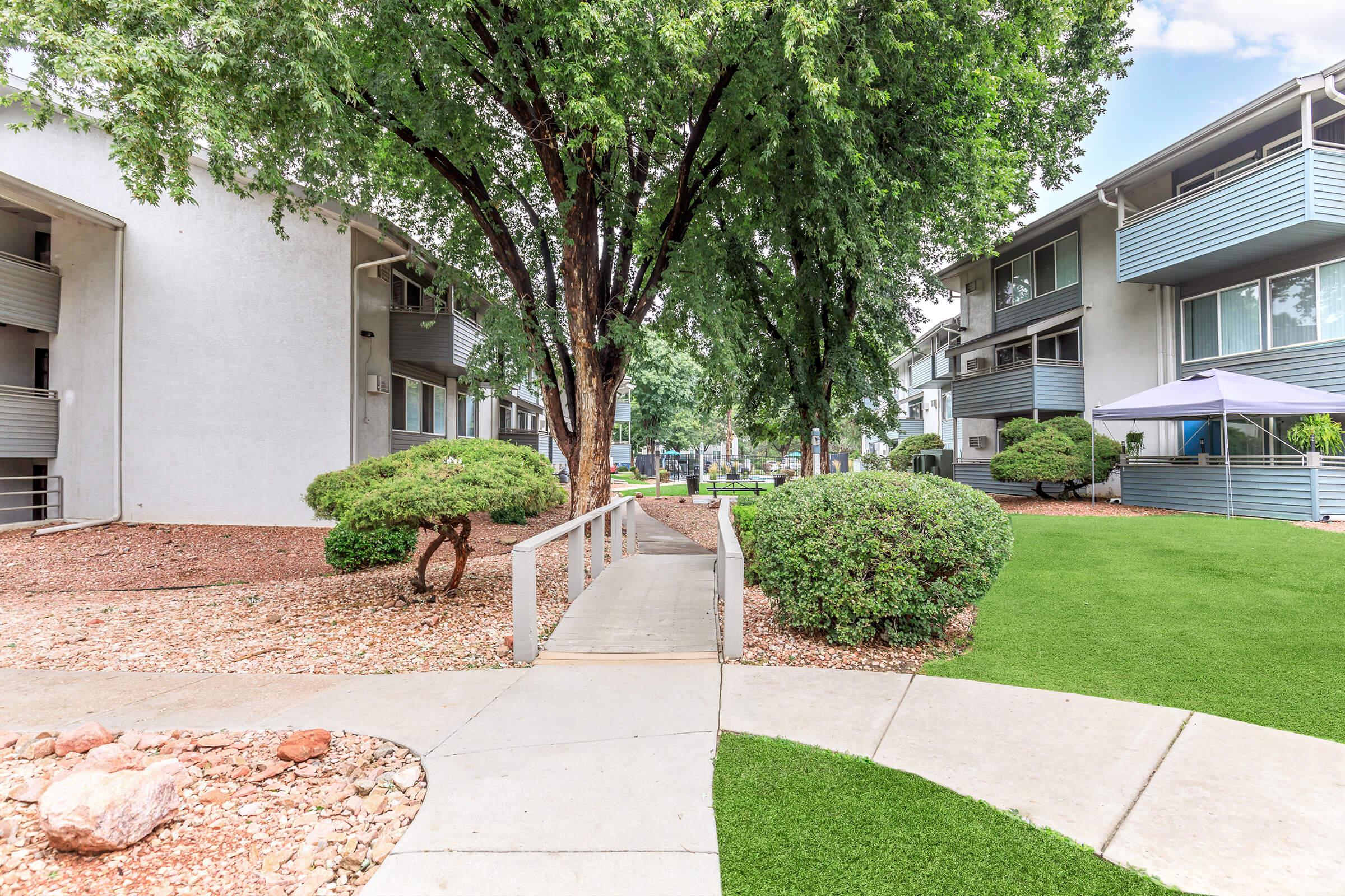 A pathway leading through a landscaped area with green grass and shrubs, flanked by two apartment buildings. Trees provide shade along the walkway, creating a welcoming atmosphere. The scene is bright and well-maintained, suggesting a comfortable living environment.