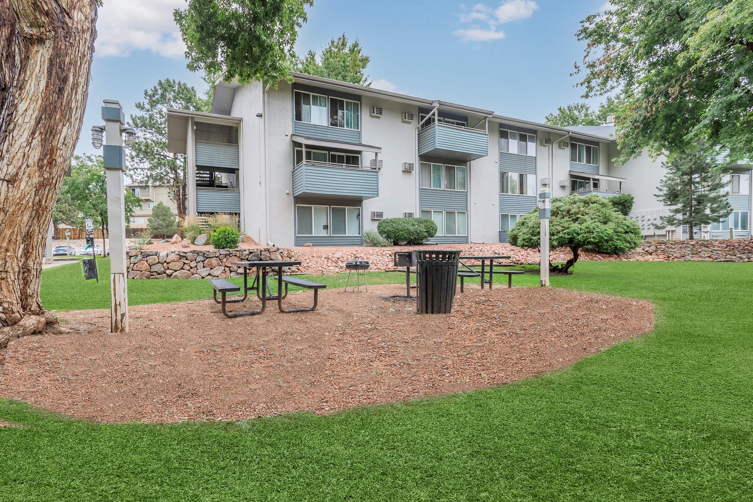A landscaped area with artificial grass featuring several black picnic tables and a trash bin. In the background, there is a multi-story apartment building with balconies and greenery. Decorative rocks and trees enhance the outdoor space.