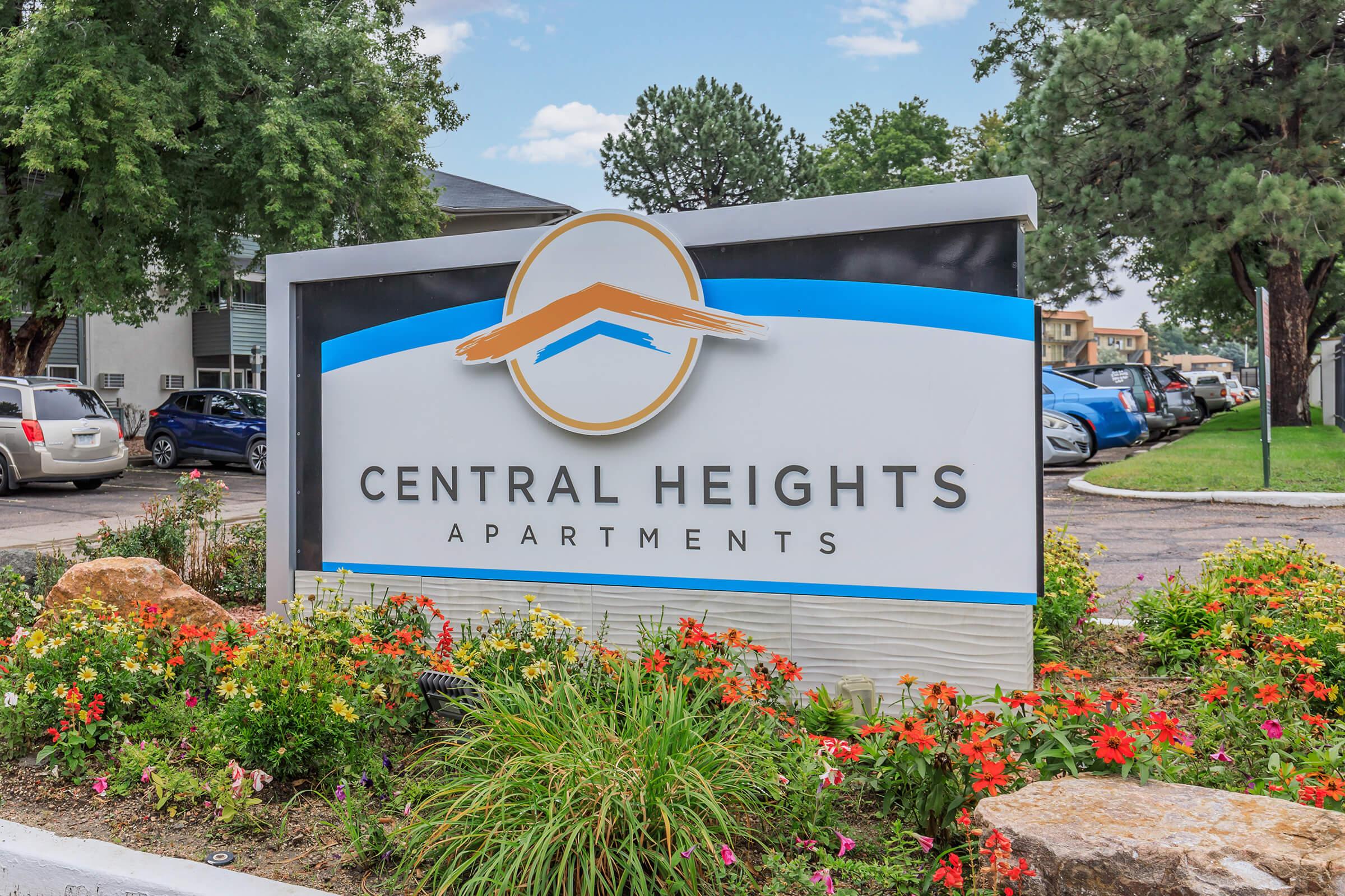 Sign for Central Heights Apartments featuring a simple logo with a mountain silhouette and decorative flowers in front. The background includes trees and parked cars, indicating a residential setting.