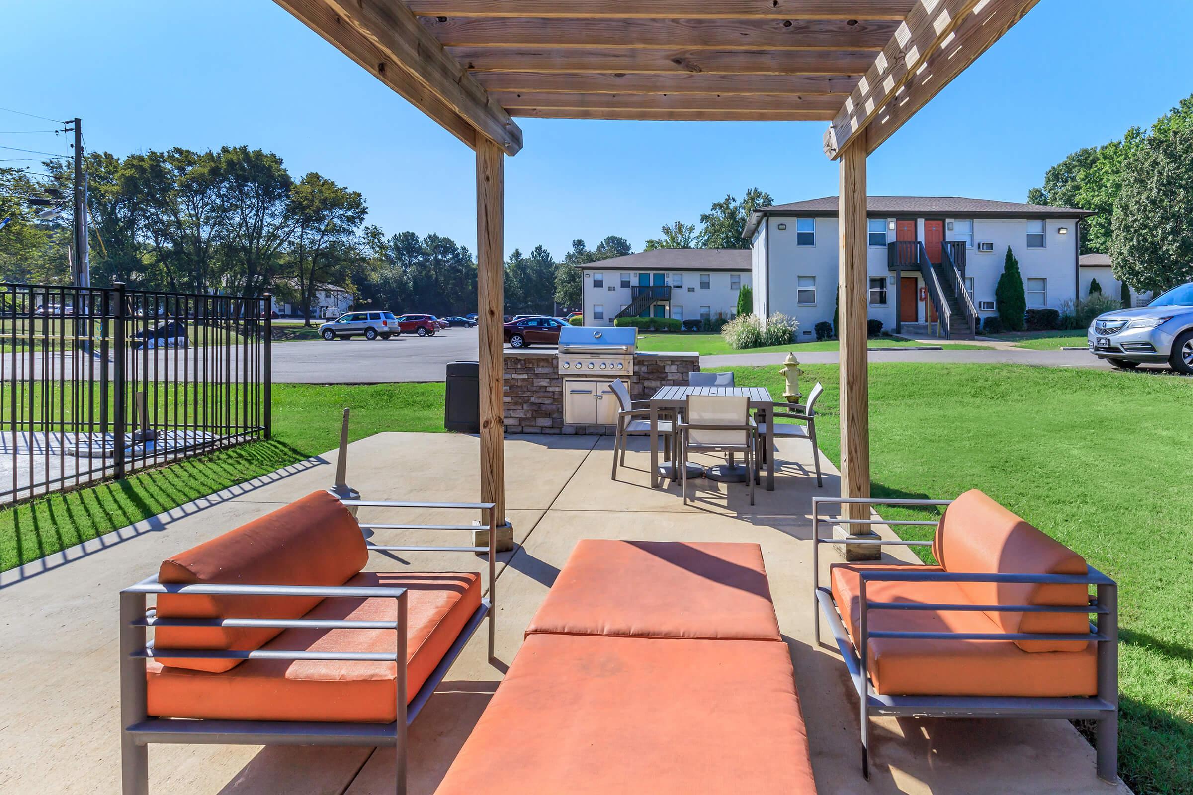 Outdoor patio area featuring two orange lounge chairs and a dining set under a wooden pergola. In the background, there's a gas grill and a view of a parking area with multiple cars. Lush green grass surrounds the space, creating a relaxing atmosphere.