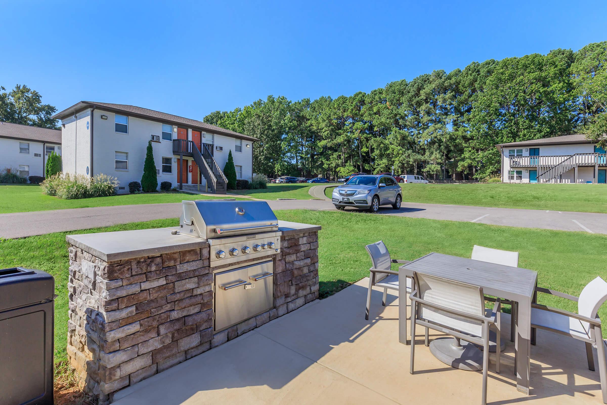 An outdoor area featuring a built-in barbecue grill made of stone, a modern dining table with chairs, and a view of an apartment building with a parking lot. Lush green trees provide background, and a car is parked on the road. Clear blue sky enhances the inviting atmosphere.