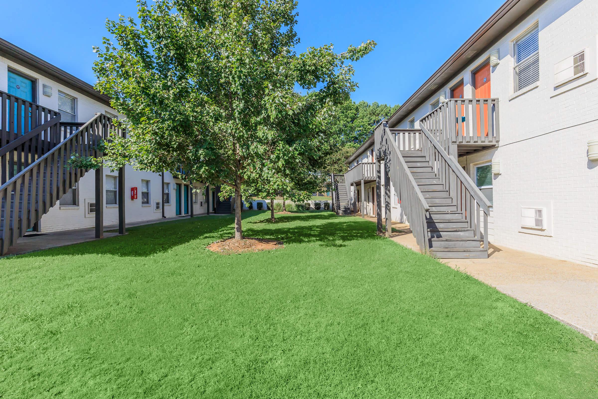 A well-maintained courtyard of an apartment complex, featuring a green lawn and a central tree. Surrounding the lawn are two sets of two-story buildings with stairs leading to individual apartments. The sky is clear blue, creating a bright and inviting atmosphere.
