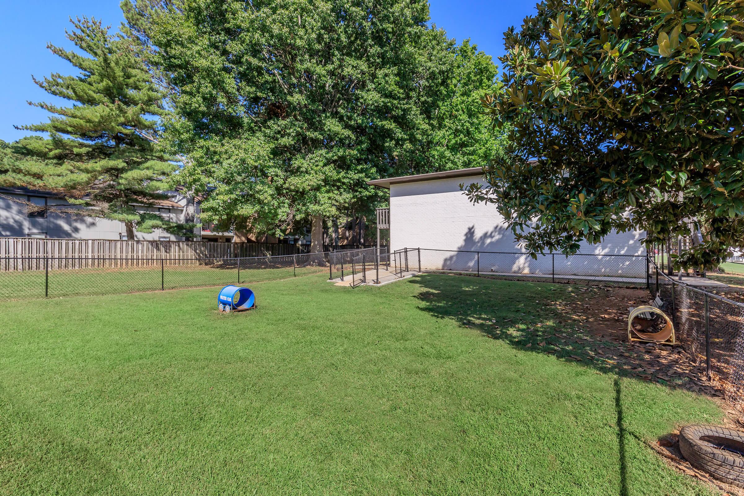A grassy yard enclosed by a fence, featuring a small blue doghouse and a yellow structure. Nearby, a white building stands amongst trees, providing shade to the area. The scene is clear and sunny, suggesting a well-maintained outdoor space suitable for pets.