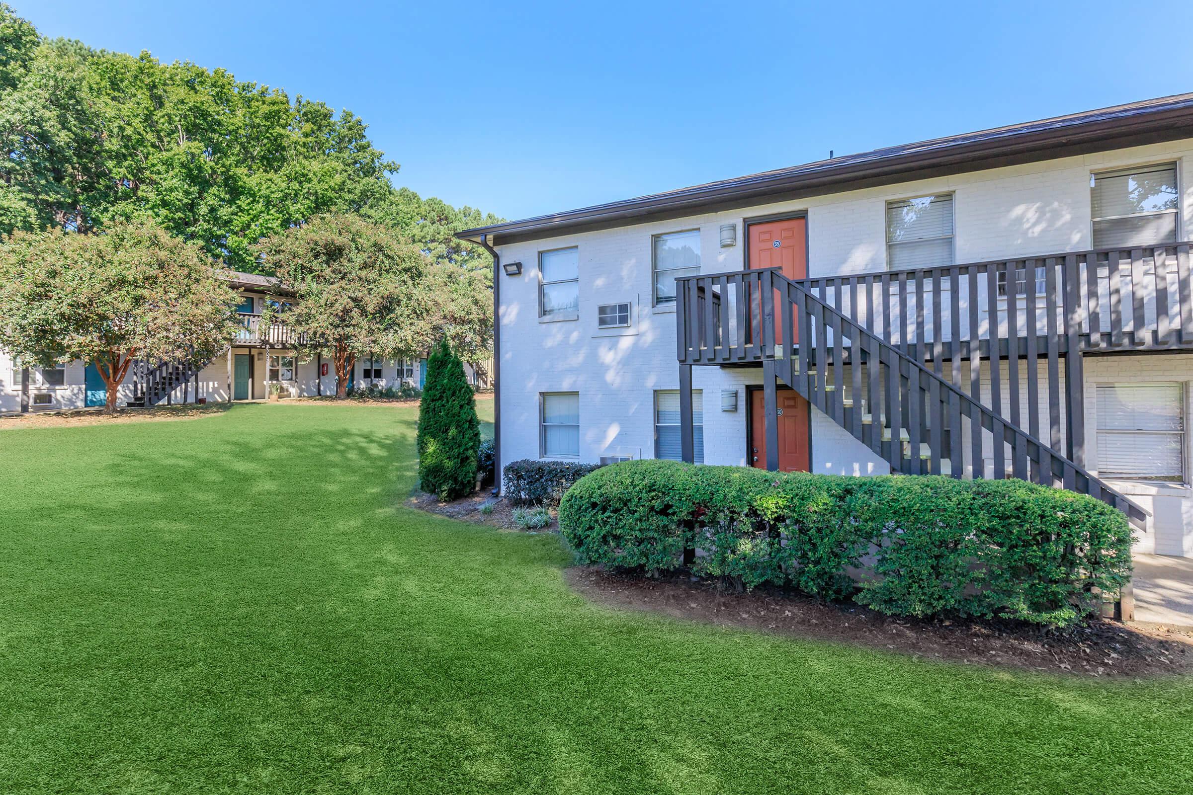 Two-story apartment building with a white exterior and orange doors, surrounded by a well-maintained lawn. There are shrubs in the foreground and trees in the background, creating a pleasant outdoor setting. The sky is clear and blue. A walkway and another building are visible in the distance.