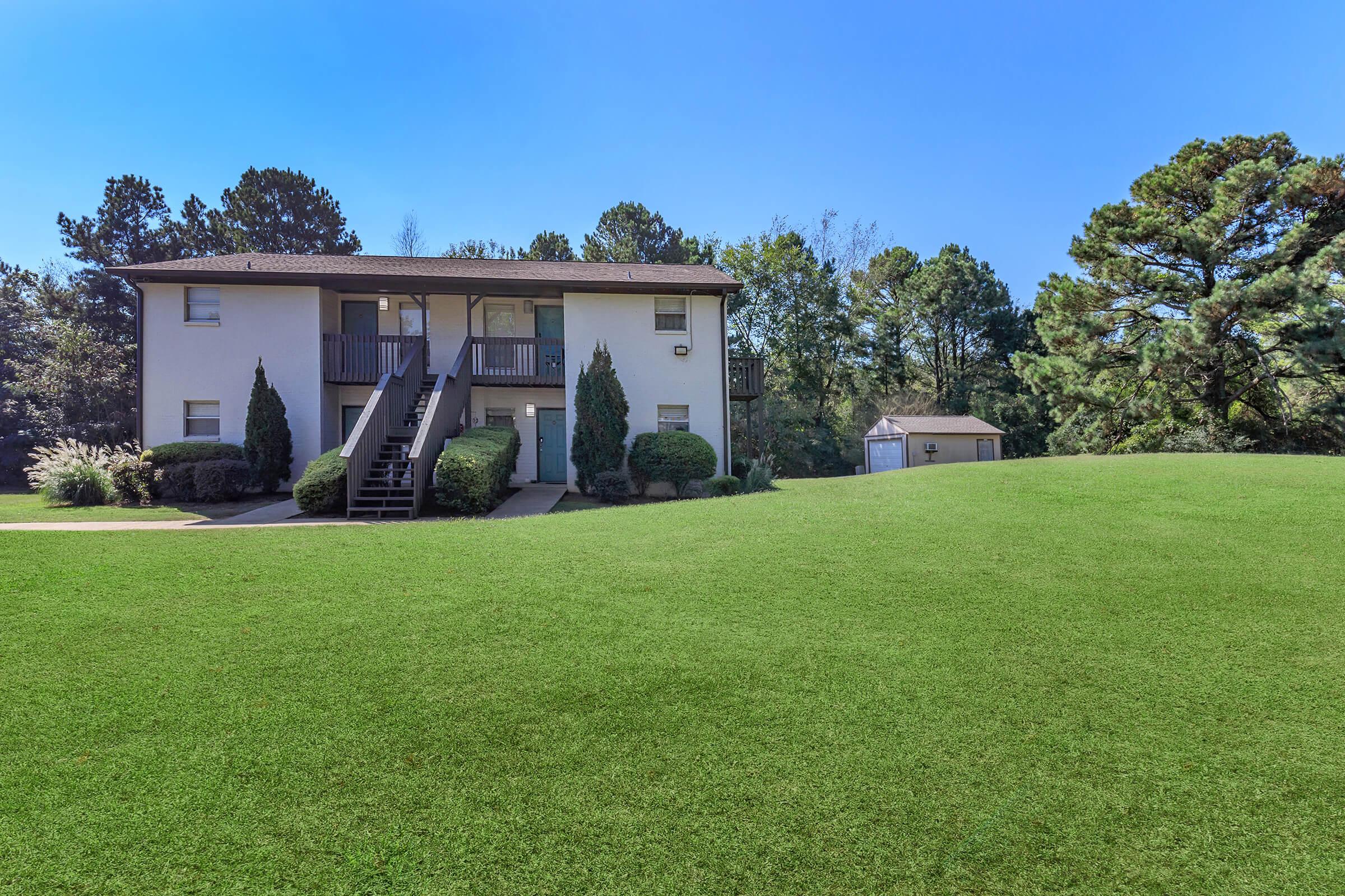 Two-story residential building surrounded by a well-manicured lawn, with a gravel path leading to the entrance. The landscape features green bushes and trees in the background under a clear blue sky. A small shed is visible to the right of the building.