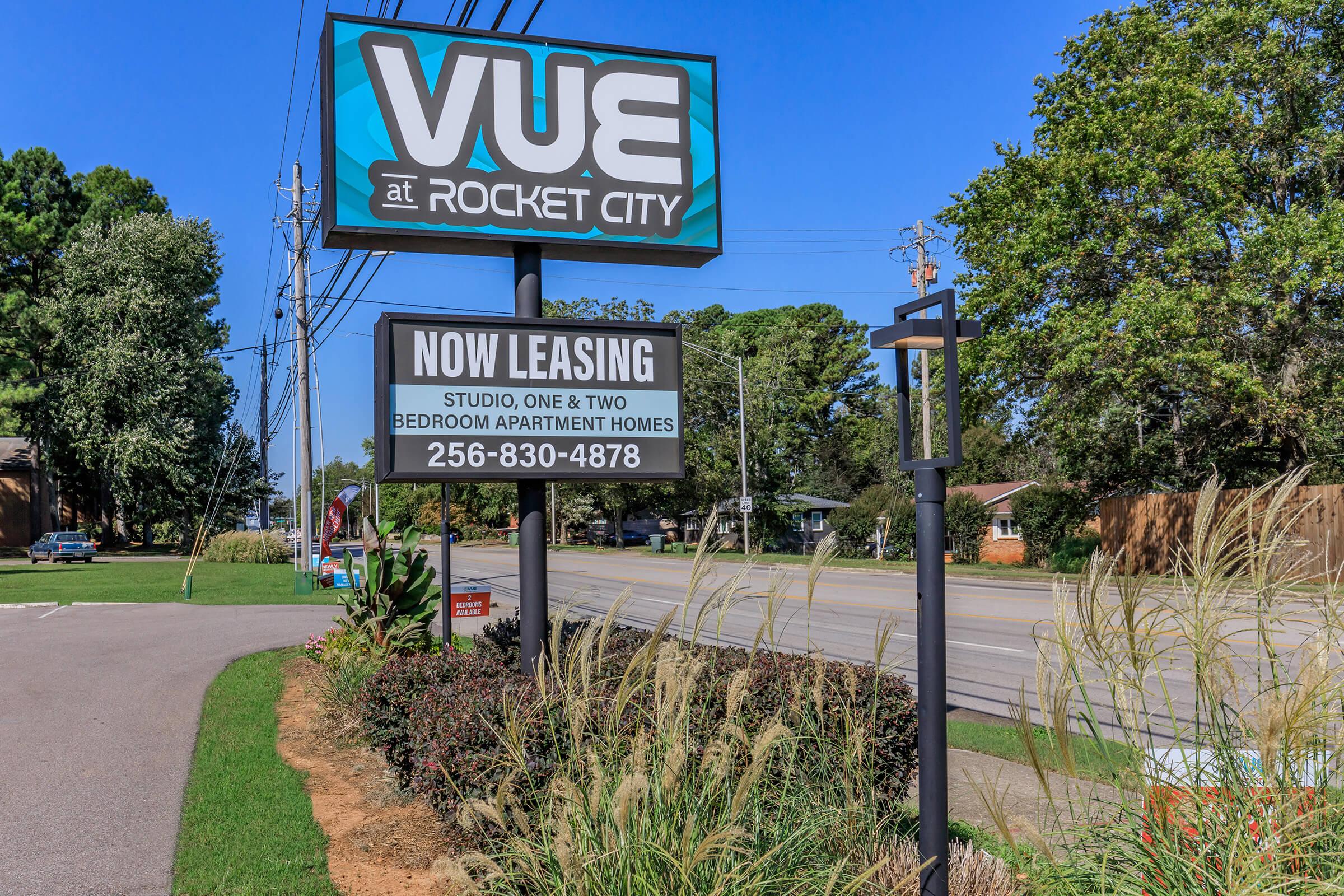 A sign for "Vue at Rocket City" advertising apartment homes. It mentions "Now Leasing" for studio, one, and two-bedroom apartments, along with a contact number: 256-830-4878. The sign is surrounded by greenery and is situated near a road on a sunny day.