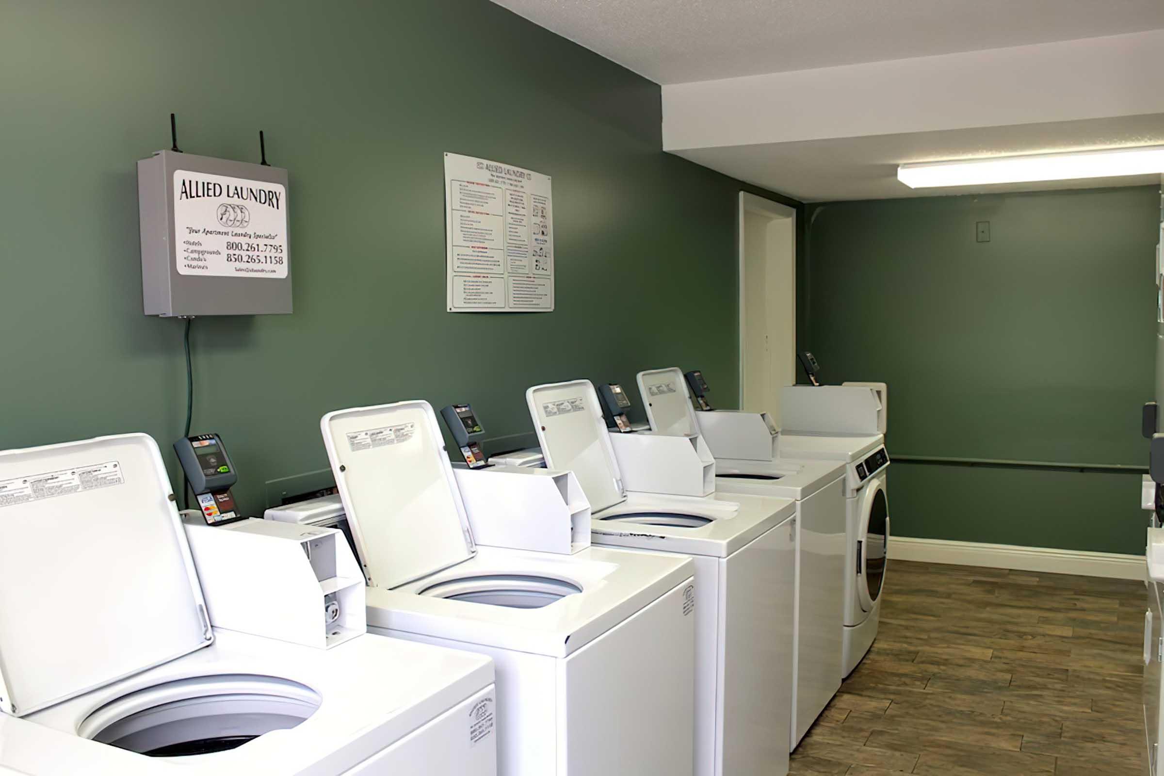 A clean and well-lit laundry room featuring several white washing machines lined up against a green wall. A bulletin board and informational sign are mounted on the wall, and the floor is covered with wood-like tiles. The space is organized and functional for laundry use.