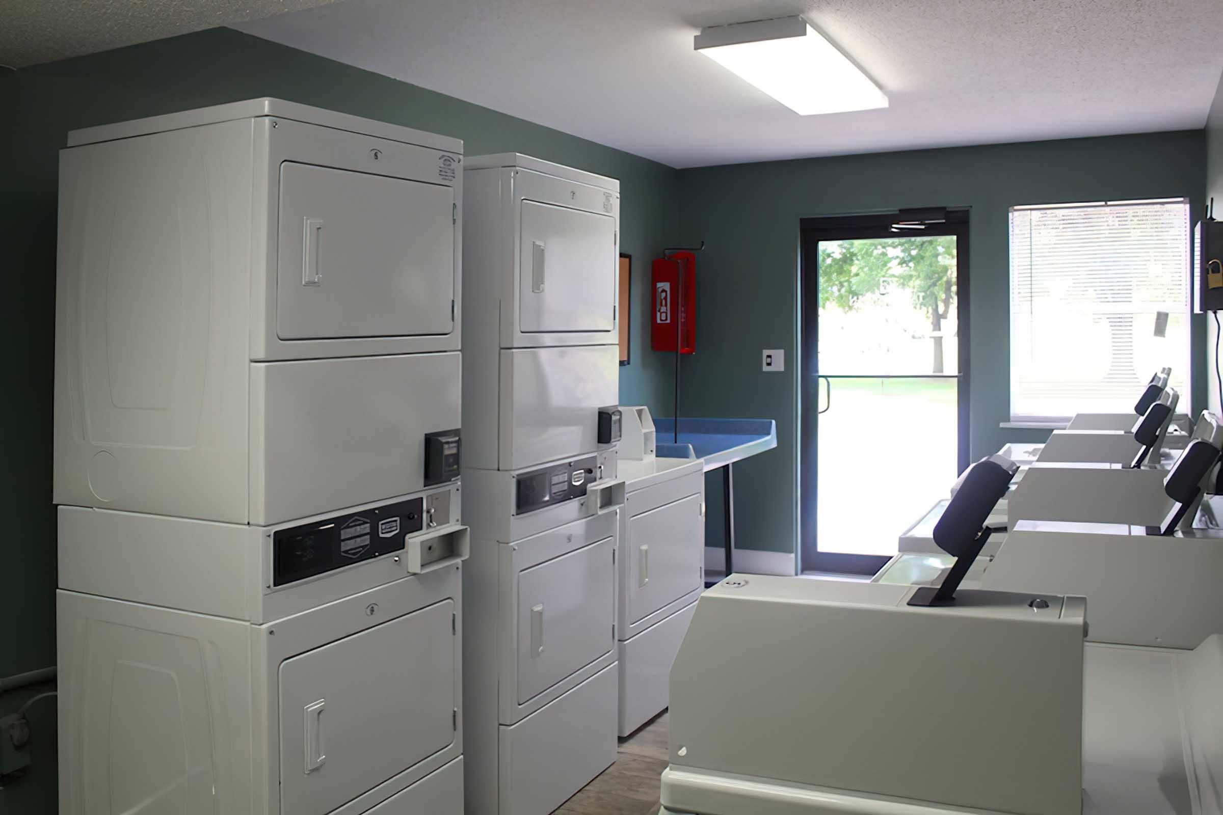 A laundromat interior featuring several white washing machines and dryers, a countertop, and a large window letting in natural light. The walls are painted a soft green, and there is a fire extinguisher mounted on the wall. The space is clean and well-lit, designed for laundry use.