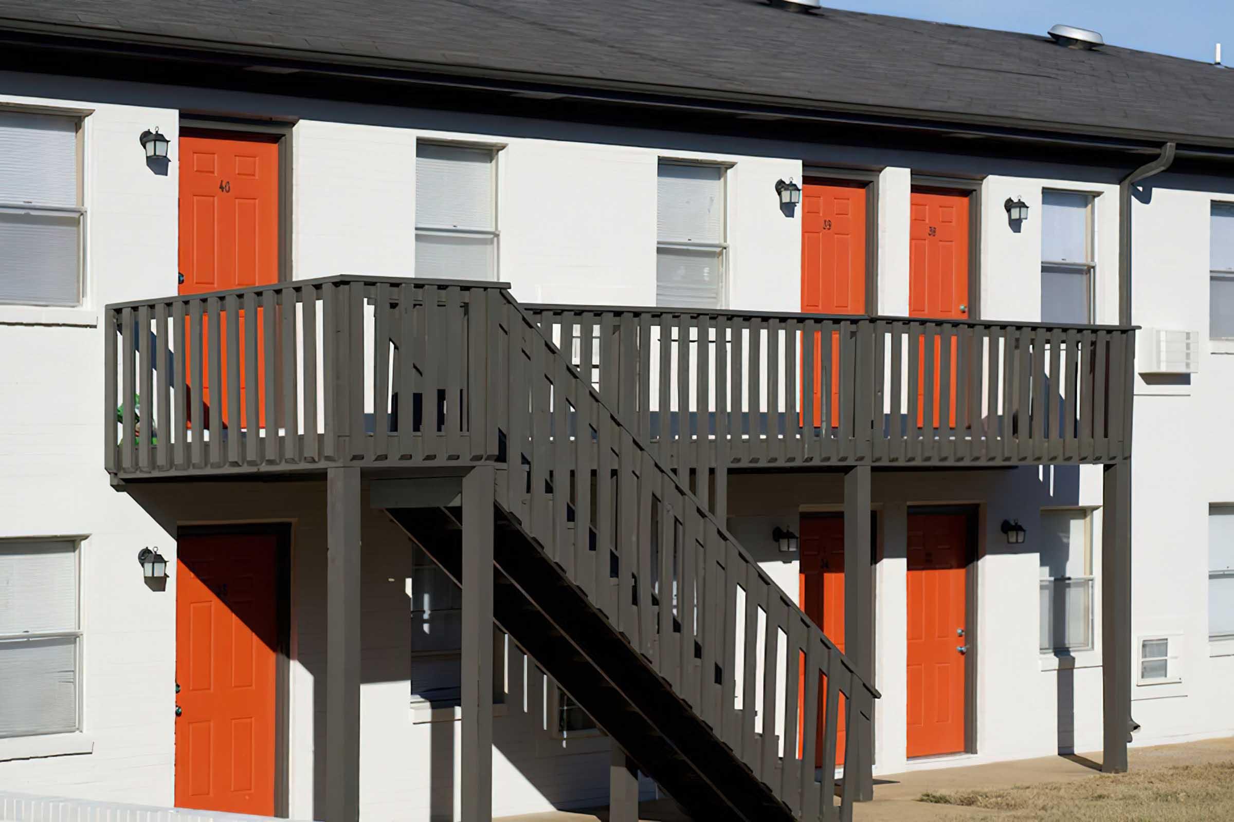 A two-story building with a white exterior featuring multiple orange doors. A dark wooden staircase leads to a balcony that connects the upper level doors. There are windows with blinds beside each door and wall-mounted light fixtures. The setting appears to be a residential area.