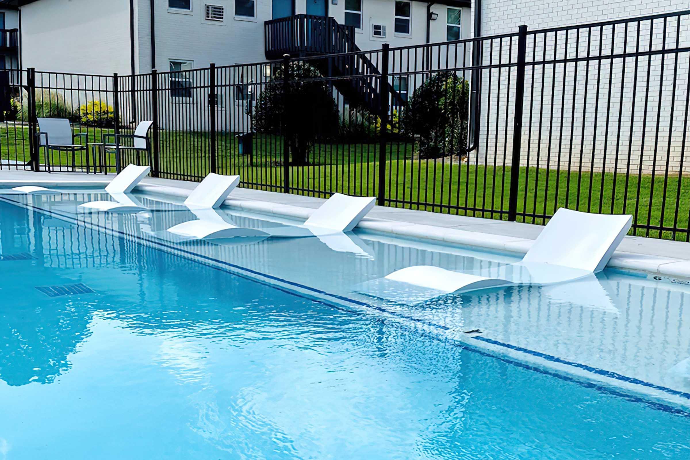 A swimming pool with shallow water featuring modern white lounge chairs partially submerged along the edge. In the background, a well-maintained lawn and a building with balconies are visible, enclosed by a black fence. Clear blue sky reflects on the water surface.