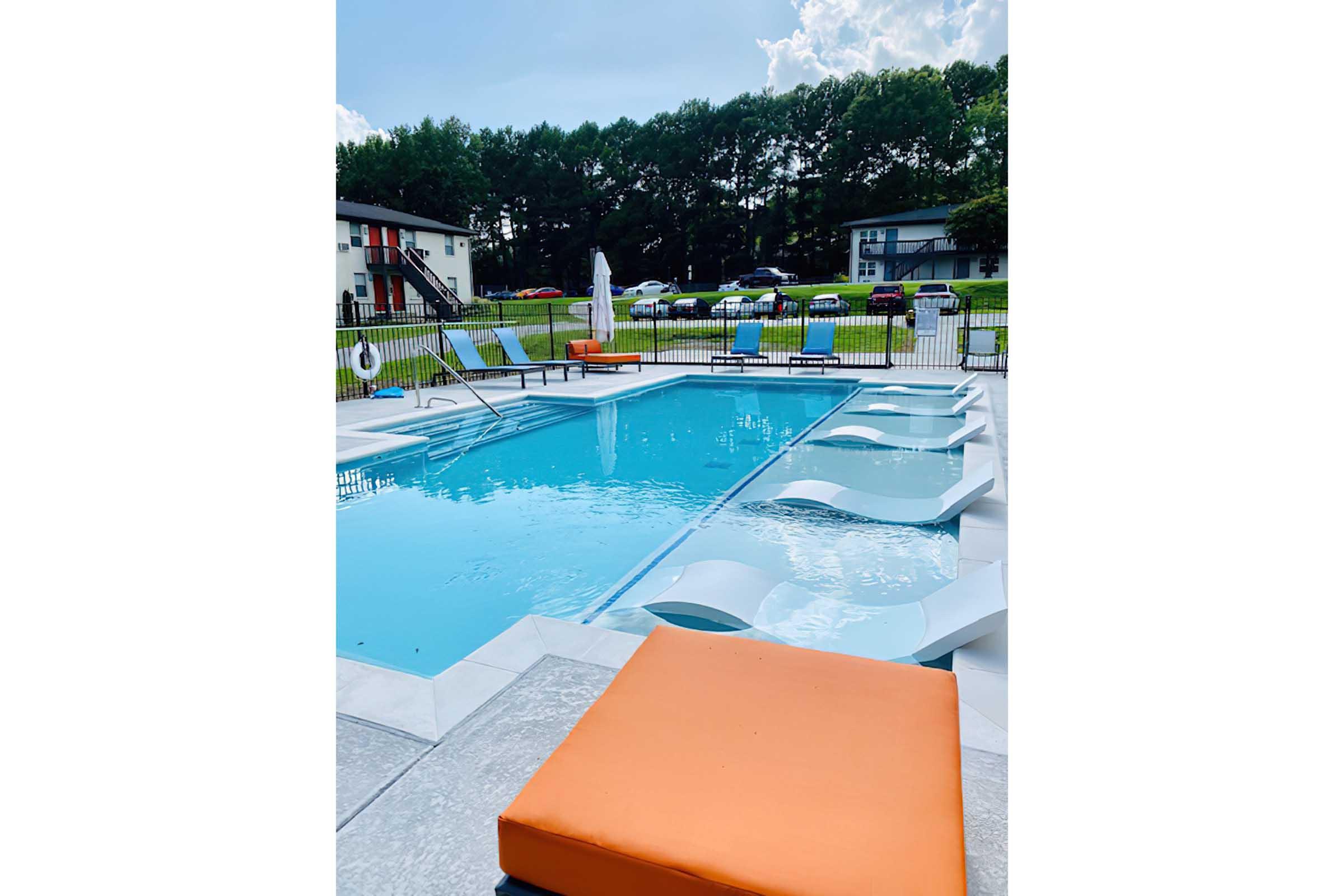 A clear swimming pool surrounded by a patio area featuring orange lounge chairs. There are a few poolside loungers partially submerged in the water. In the background, there are green trees and a couple of buildings visible, along with parked cars on a sunny day.