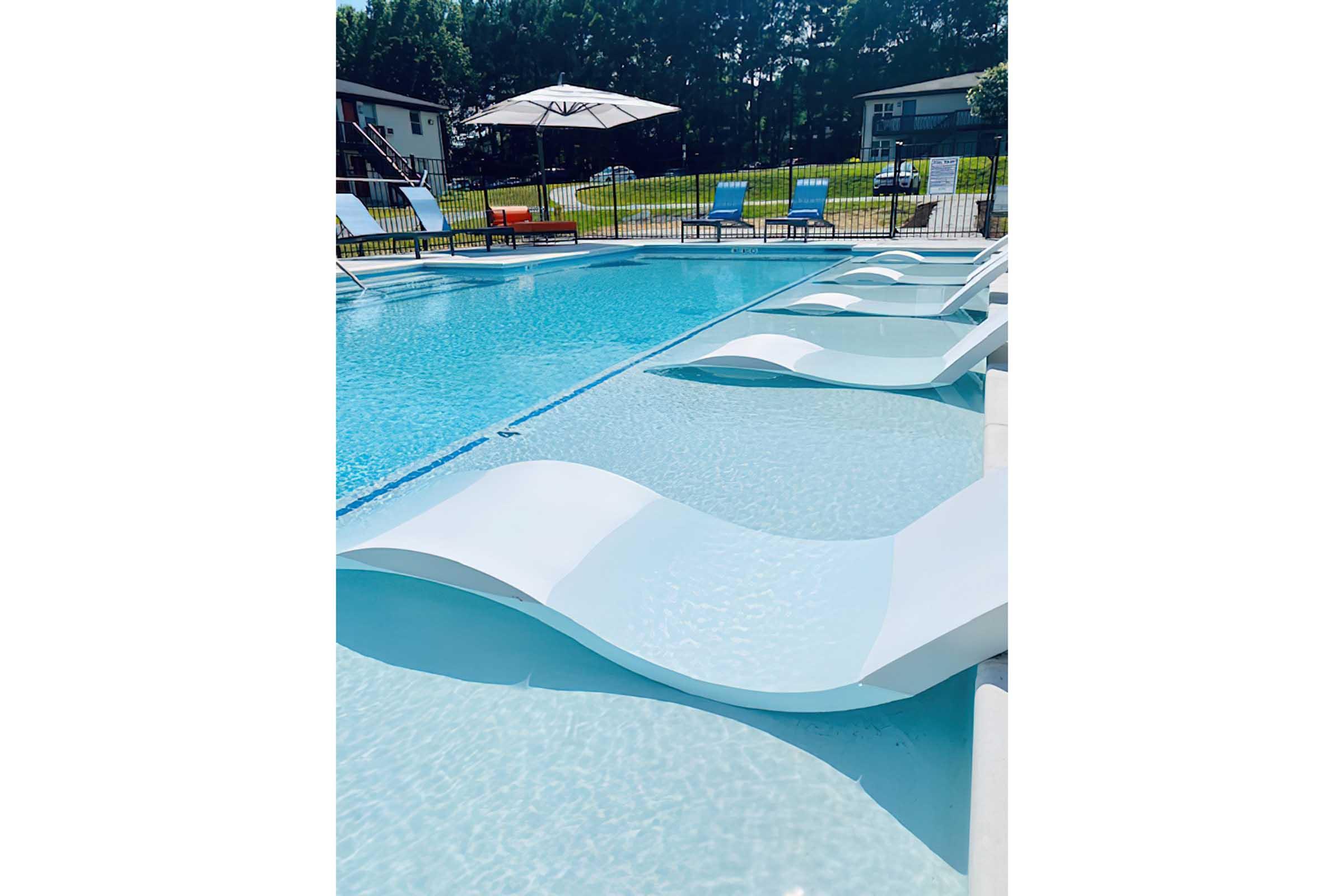 A tranquil pool area featuring a blue swimming pool with stylish white loungers partially submerged in the water. Bright sun loungers in various colors are arranged around the pool, with a large umbrella providing shade. Lush greenery is visible in the background, creating a serene atmosphere.