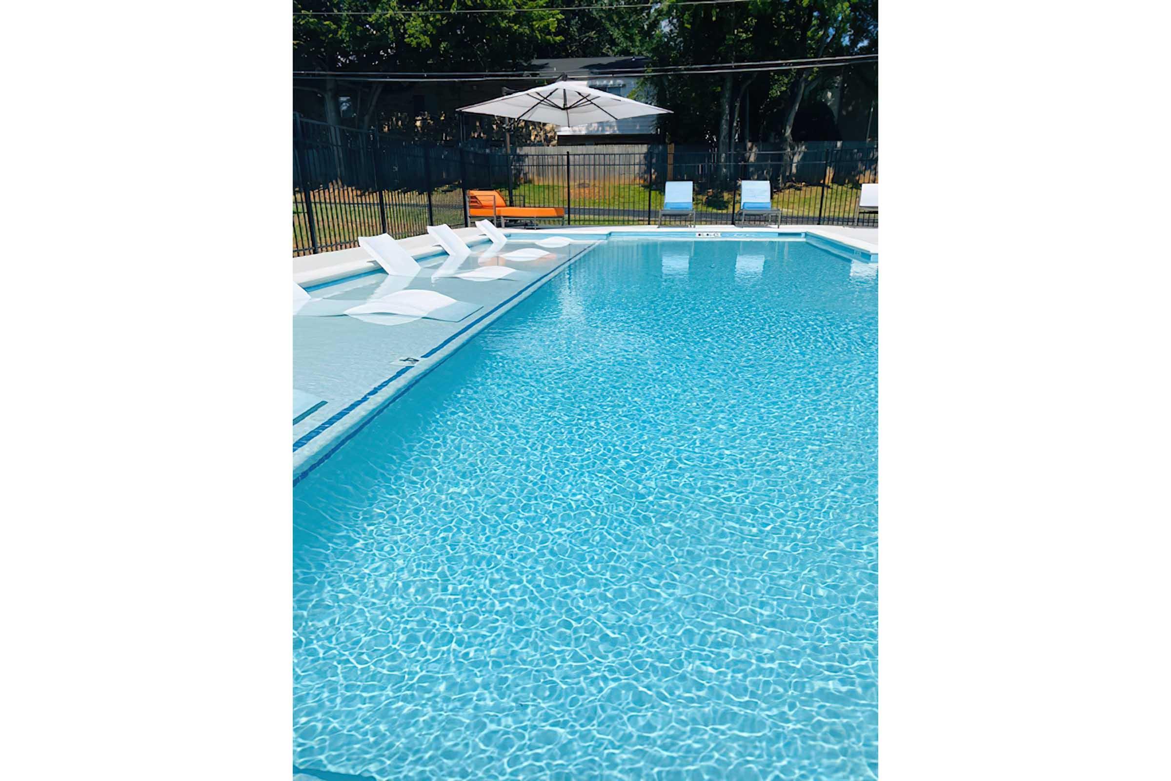 A bright blue swimming pool with clear water, surrounded by a fence. White lounge chairs are positioned along one side of the pool, and a large umbrella provides shade over the seating area. Lush greenery can be seen in the background, creating a relaxing outdoor setting.