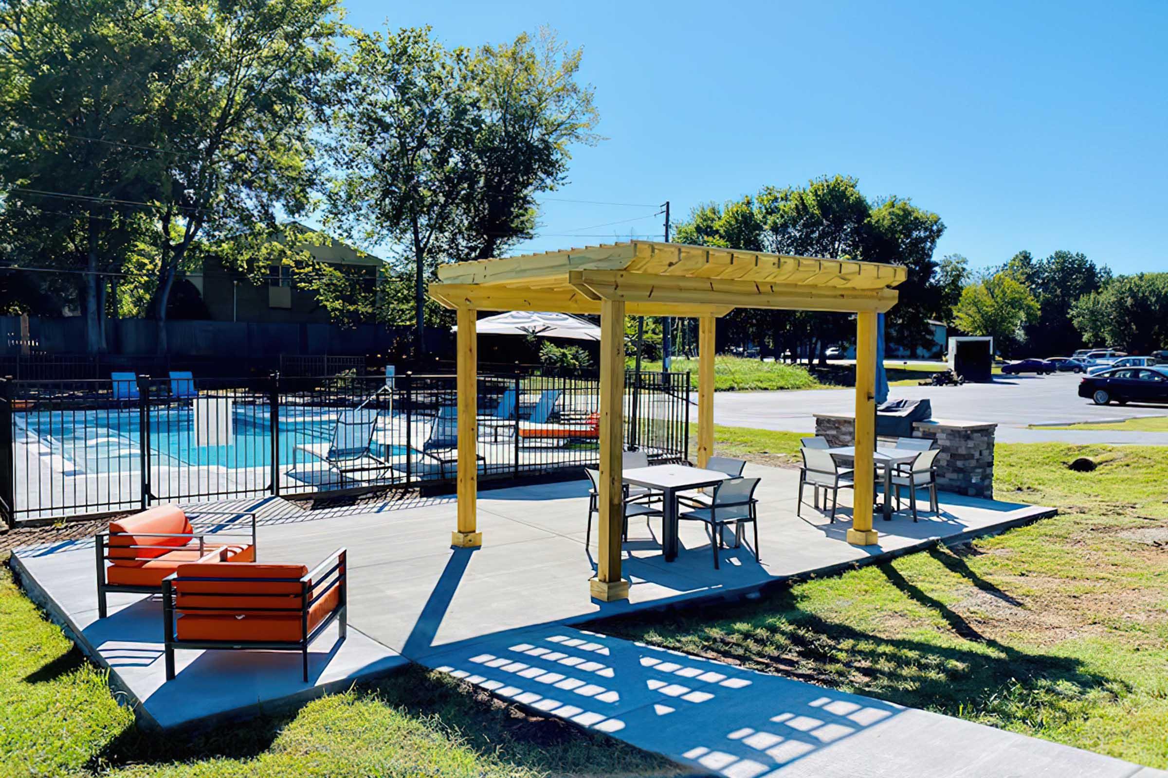A bright, sunny poolside area featuring a yellow pergola with tables and chairs. Red outdoor seating is nearby. The pool is visible in the background, surrounded by a black fence and greenery. The sky is clear and blue, creating a welcoming atmosphere for relaxation and socializing.