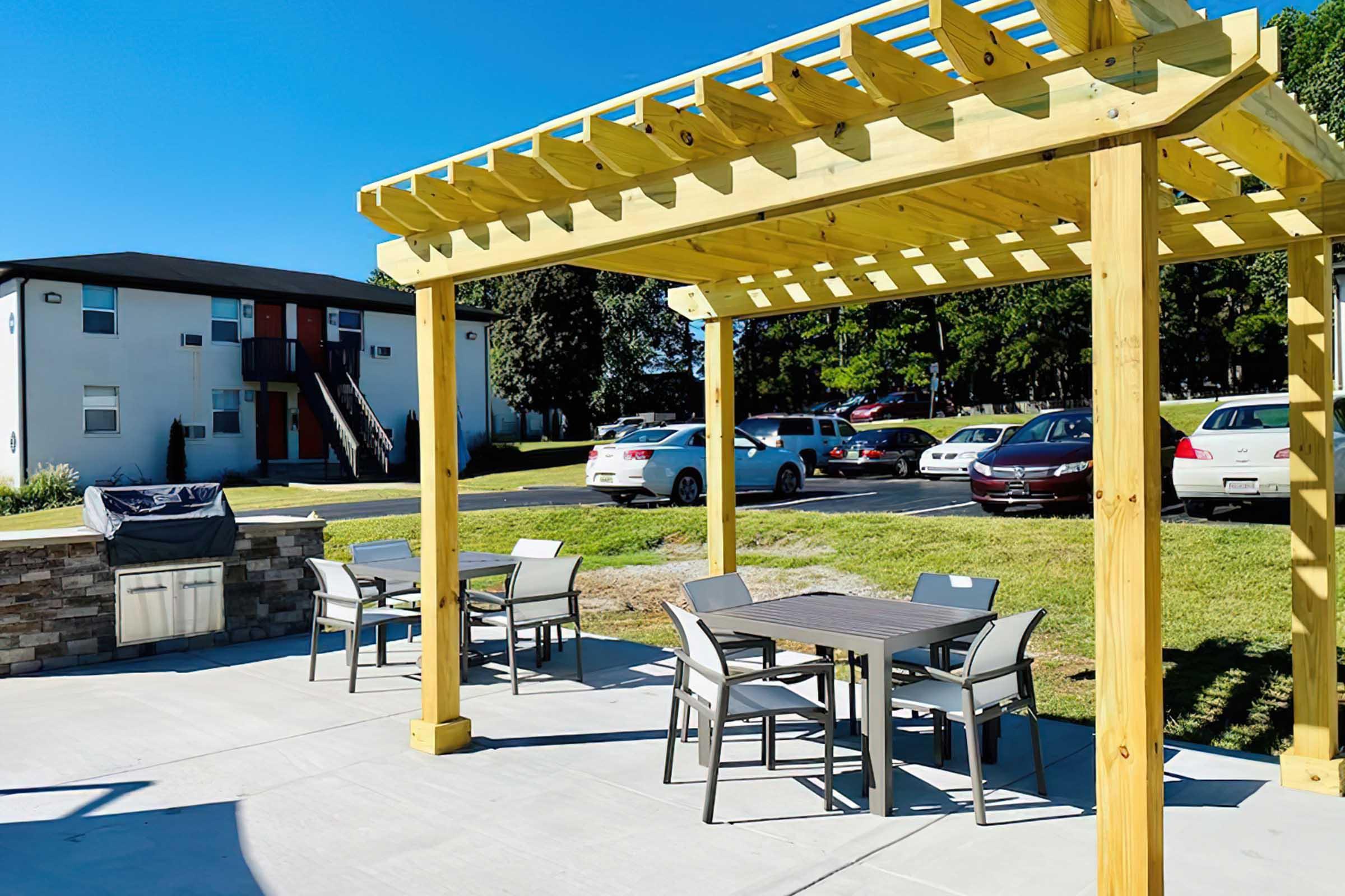 Outdoor patio area with a wooden pergola, featuring several tables and chairs. A barbecue grill is present nearby, and multiple parked cars can be seen in the background, along with an apartment building. The scene is bright and sunny, set in a green space.