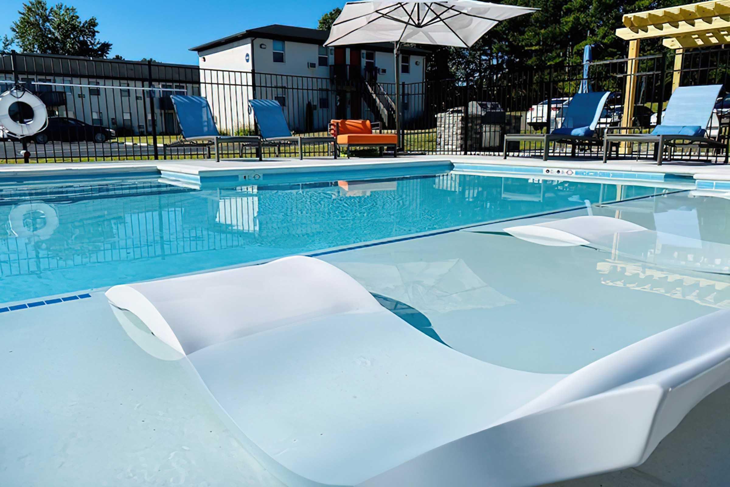 A clear swimming pool featuring white lounge chairs partially submerged in the water. In the background, there are colorful poolside chairs, a large umbrella for shade, and a fence surrounding the pool area. The scene has a bright, sunny atmosphere with green trees in the distance.