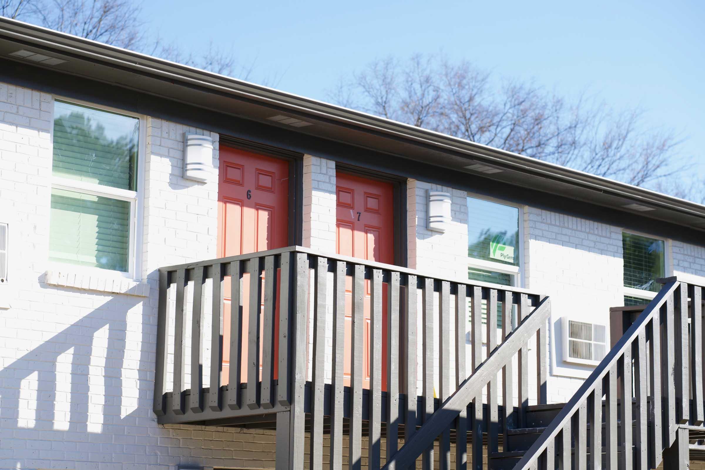 Two doors numbered 6 and 7, painted in bright orange, located on an exterior balcony of a two-story white brick building. Metal stair railing leads up to the doors, which are flanked by windows with green and blue accents. The building is set against a clear blue sky with trees in the background.