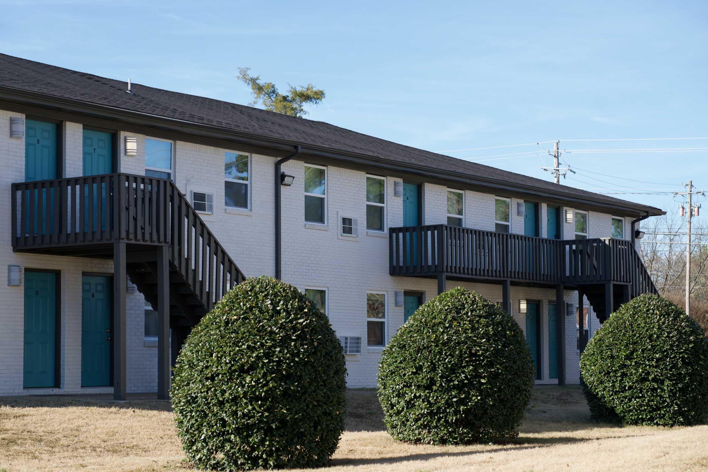 A two-story apartment building with white brick exterior and teal-blue doors. The building features two sets of stairs leading to separate entrances on the second floor, flanked by neatly trimmed round bushes in the foreground. Power lines are visible in the background against a clear blue sky.