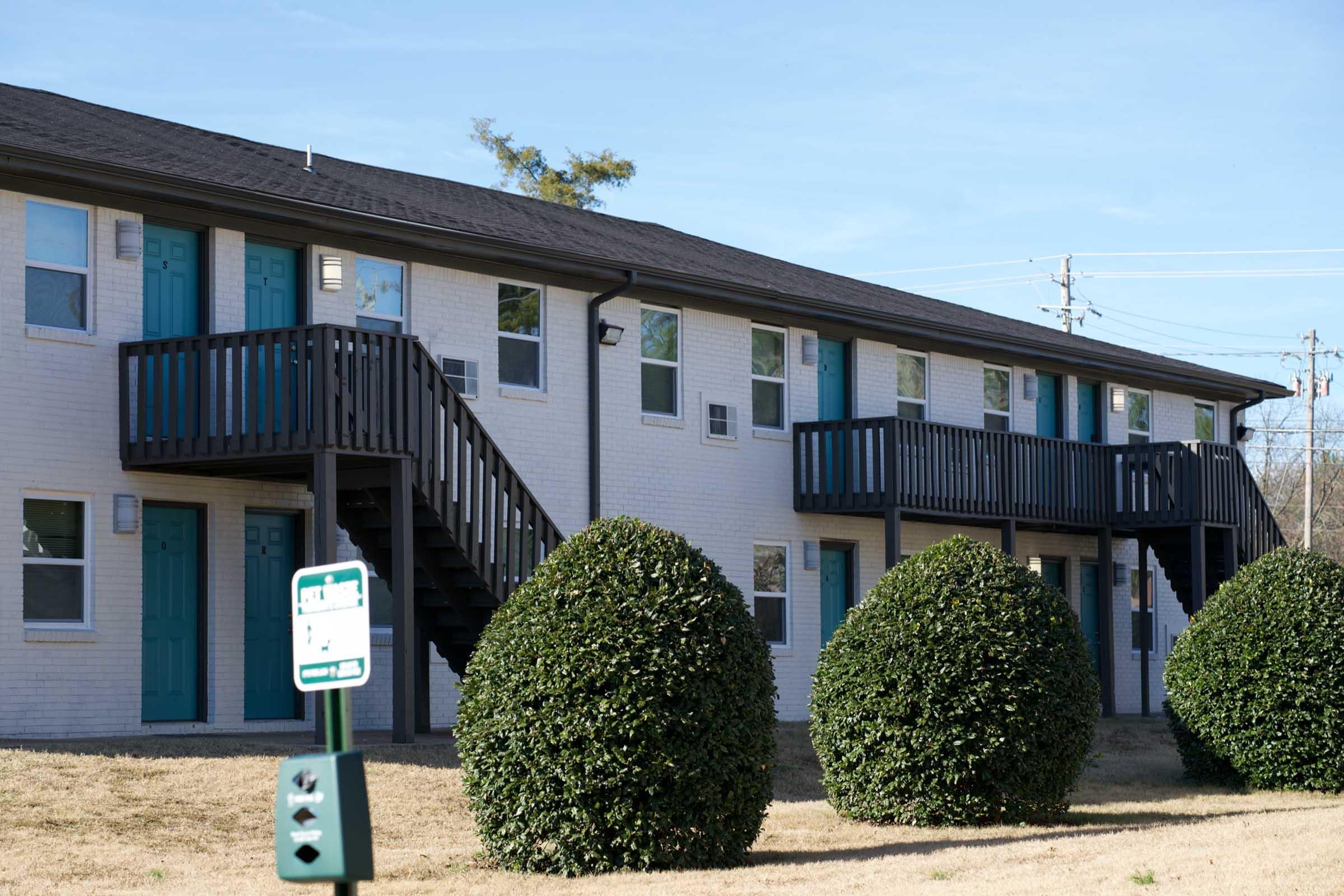 Two-story apartment building with white brick exterior and teal doors. There are two neatly trimmed round bushes in front, and a signpost is visible to the left. The sky is clear with blue hues, indicating a bright day.