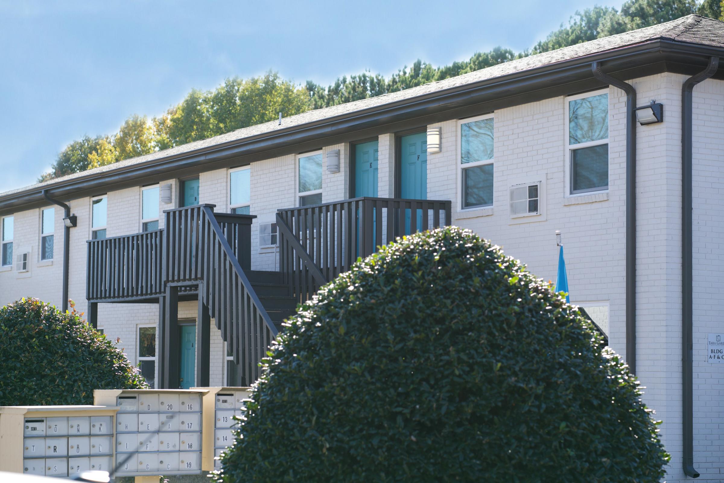 Two-story apartment building with a white brick exterior and turquoise doors. A set of black stairs leads to the upper level, flanked by trimmed bushes. In the foreground, there are mailbox units arranged neatly. The background features trees with green leaves, suggesting a sunny day.