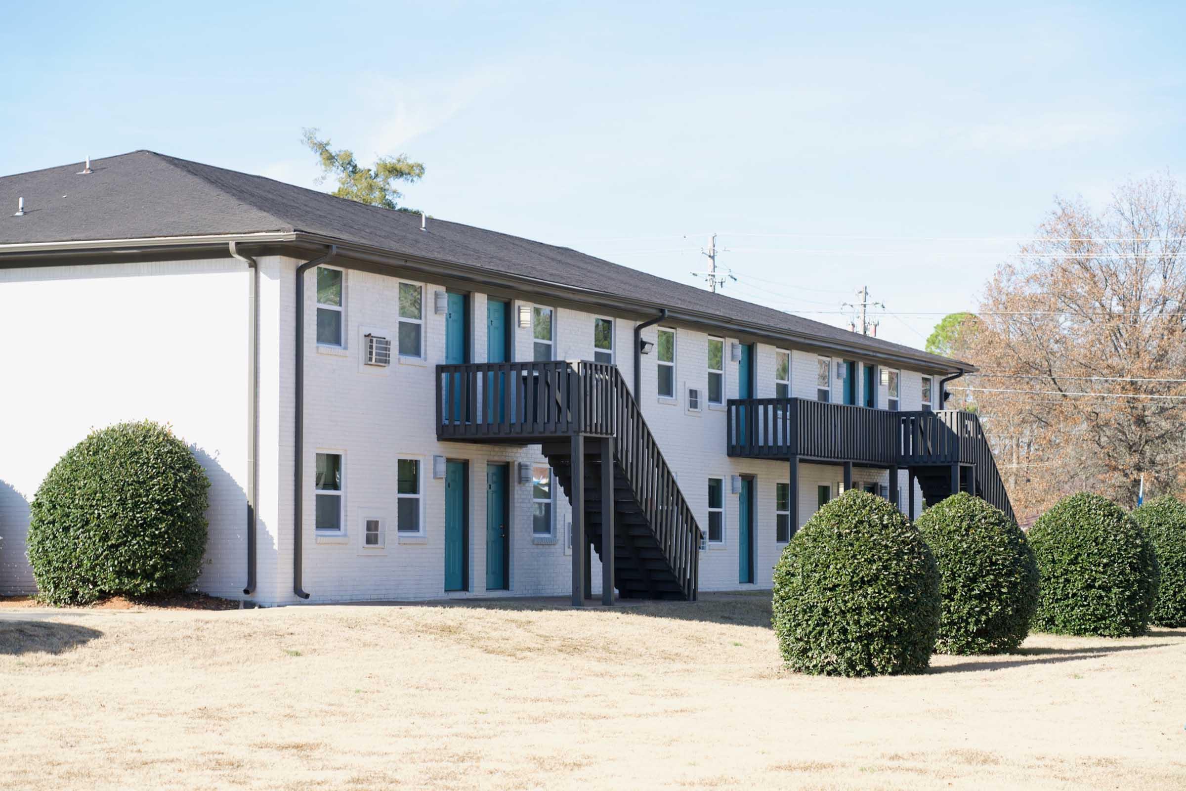 A two-story apartment building with a white exterior and blue doors. The building features outdoor staircases on both sides. Neatly trimmed bushes are planted in the foreground, and the scene is set against a clear blue sky with a few trees in the background.
