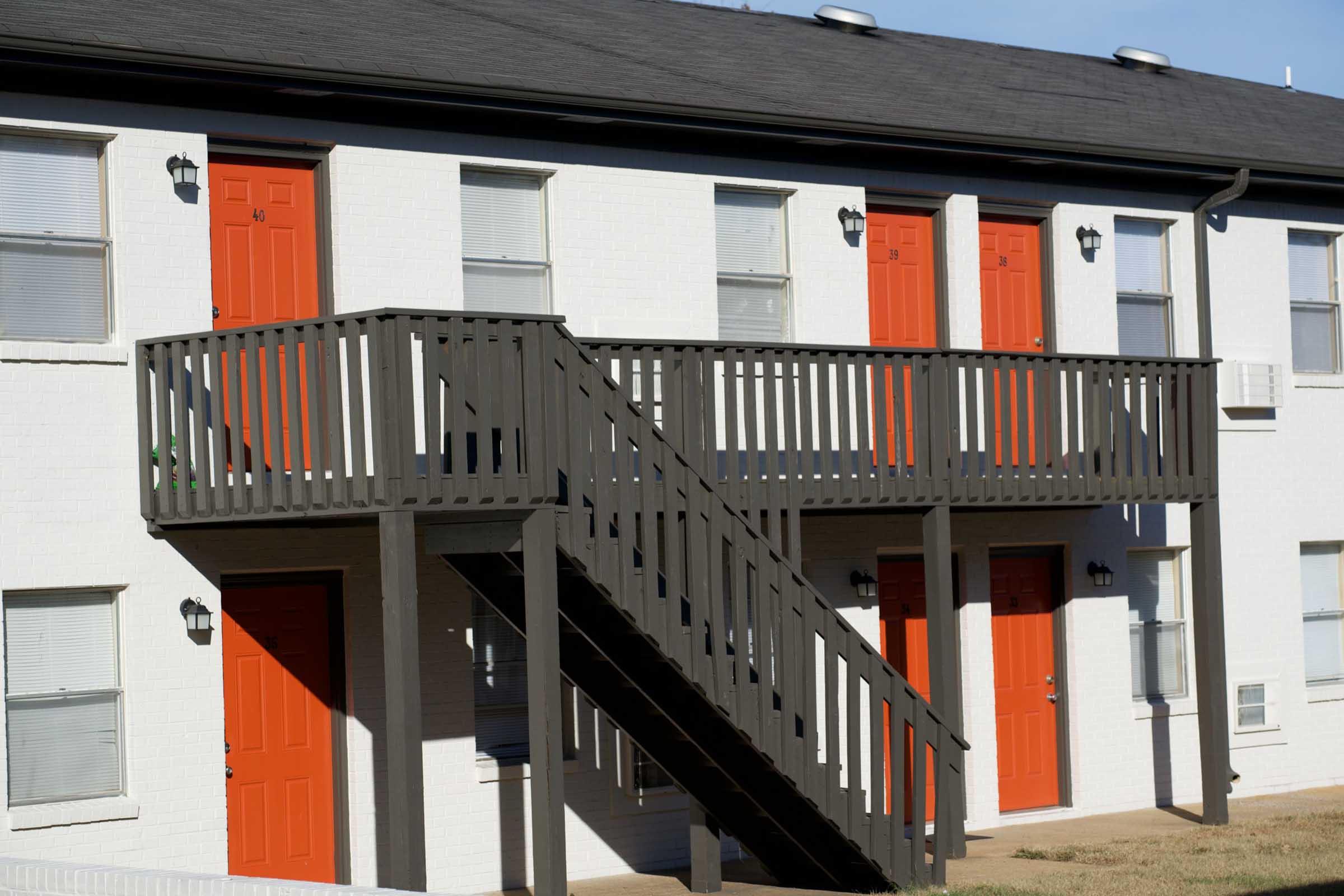 Two-story building with white exterior walls and bright orange doors. A wooden staircase leads up to a second floor balcony with a railing. Windows with blinds are visible beside the doors, and there are sconces mounted on the wall near the doors. Grass is seen in the foreground.