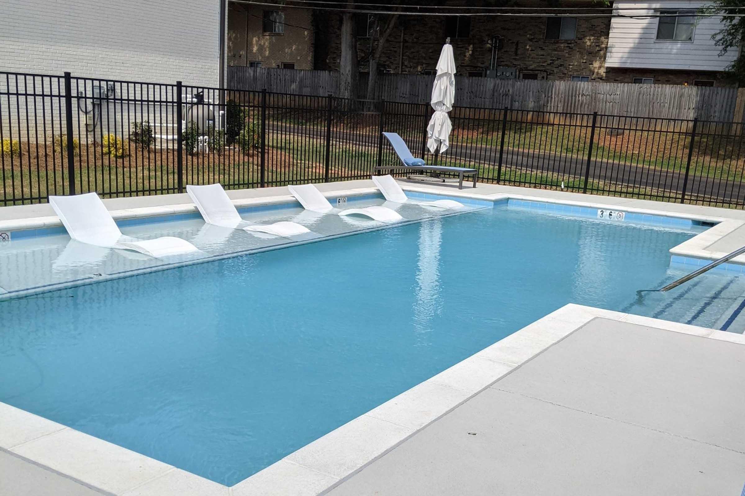 A clear swimming pool with white lounge chairs partially submerged along one side. The pool is surrounded by a black fence, with a large umbrella nearby. Lush greenery and a residential area can be seen in the background, providing a tranquil setting.