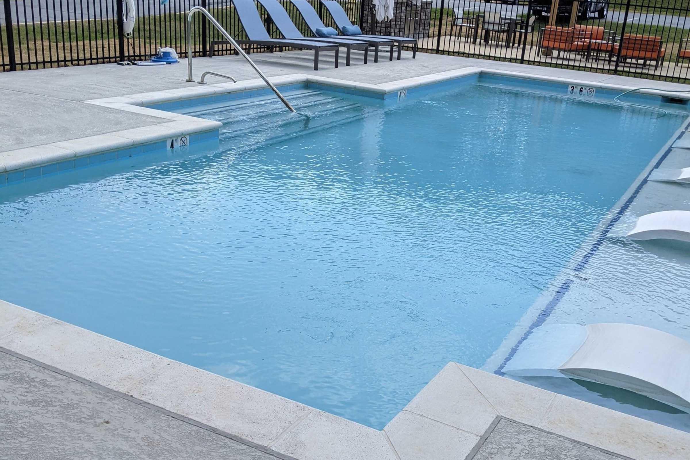 A clear swimming pool with a gentle slope, surrounded by a light-colored concrete deck. There are several lounge chairs positioned nearby, and a safety fence encloses the area. The water appears calm and inviting, reflecting the surroundings.