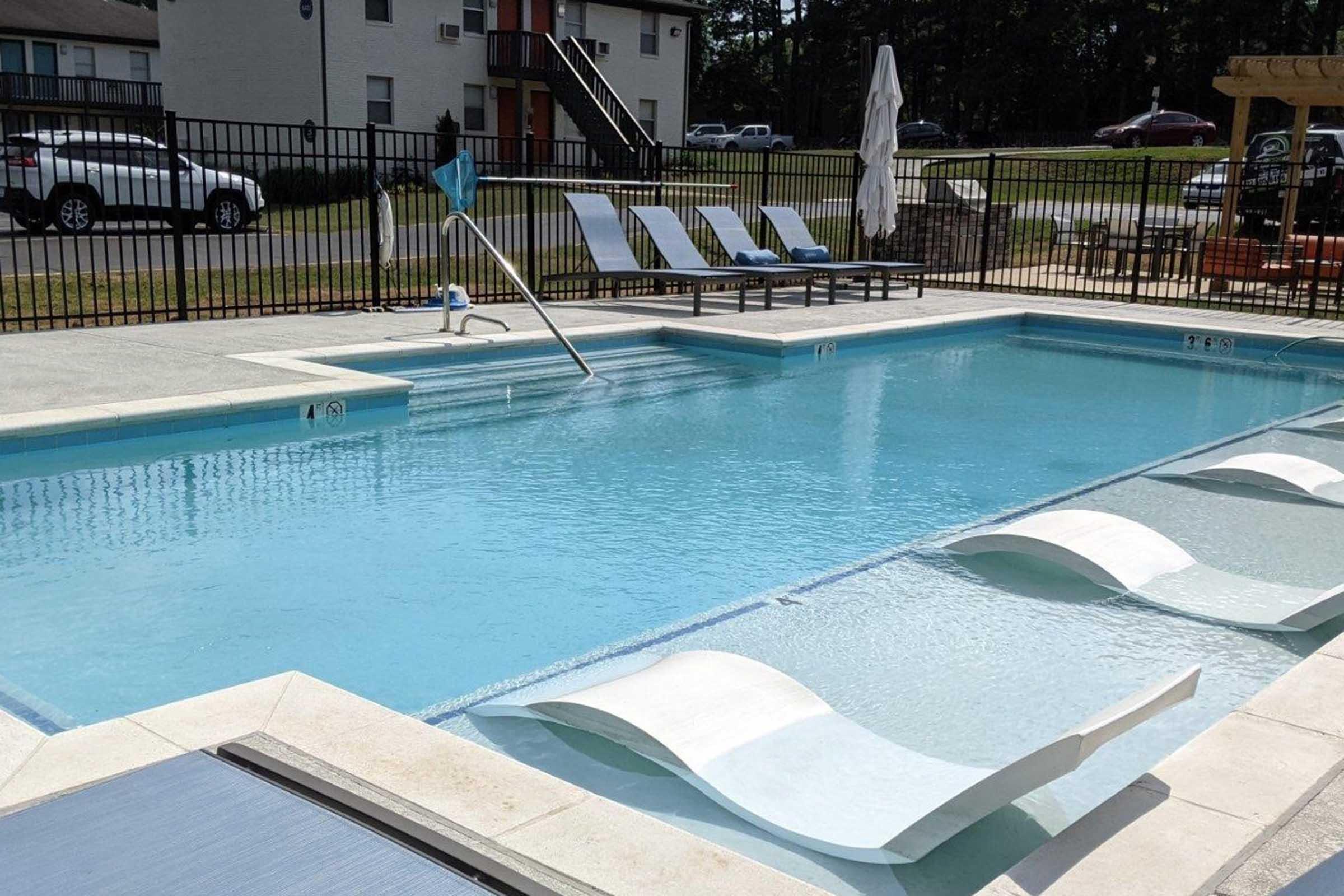 A sparkling blue swimming pool surrounded by lounge chairs. A gentle slope leads into the shallow end, and a safety handrail is positioned at the steps. In the background, there are apartment buildings and a parking area, with a few trees lining the scene.