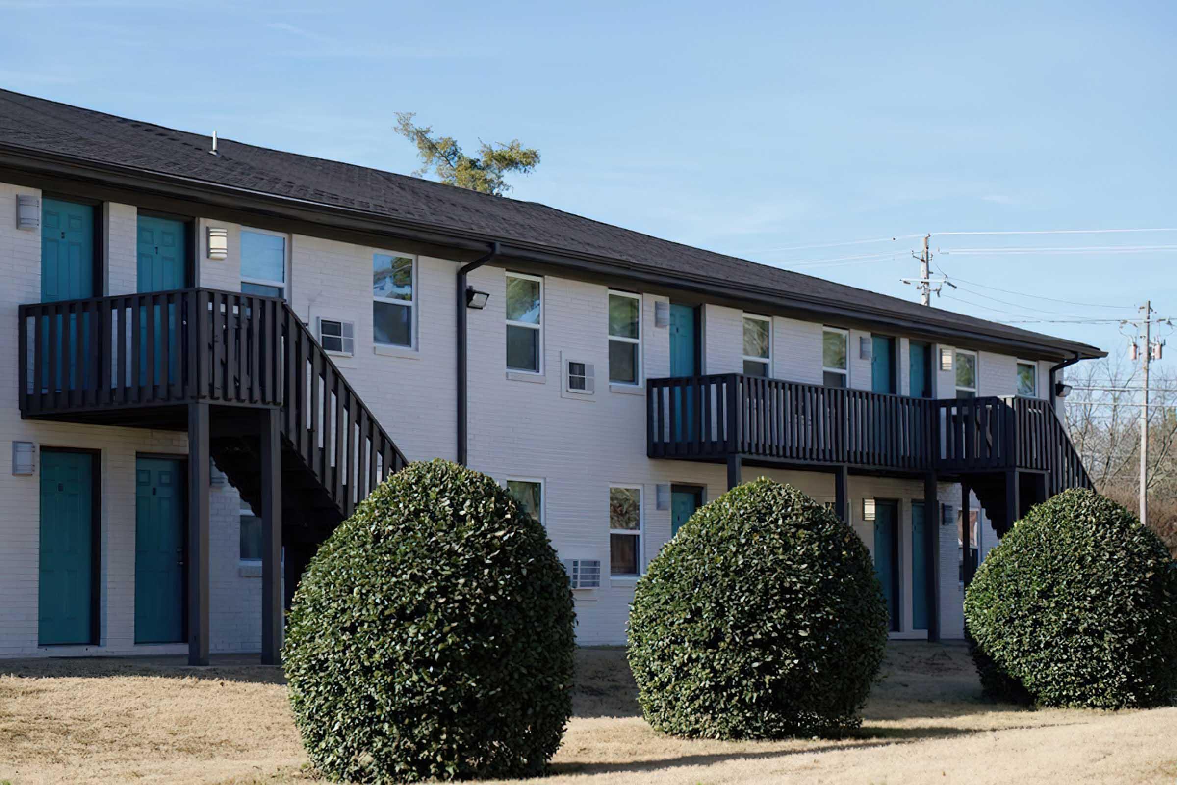 A two-story apartment building featuring multiple balconies with wooden railings. The building is painted white with teal doors. In front, there are neatly trimmed, round shrubs on grassy ground, and a clear blue sky above. Utility poles are visible in the background.