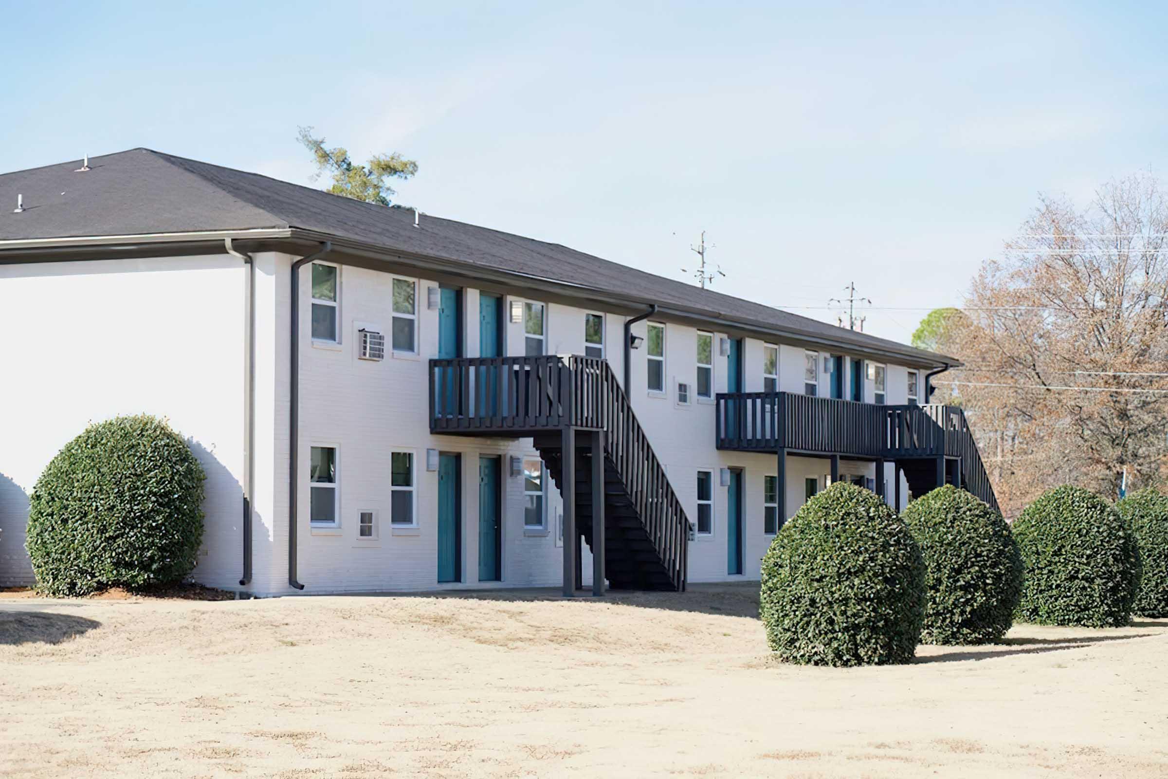 A two-story apartment building with a flat roof and balconies. The building features a mix of white and light blue façades. In the foreground, two neatly trimmed round bushes are visible on a grassy lawn, and the sky is clear with a few trees in the background.