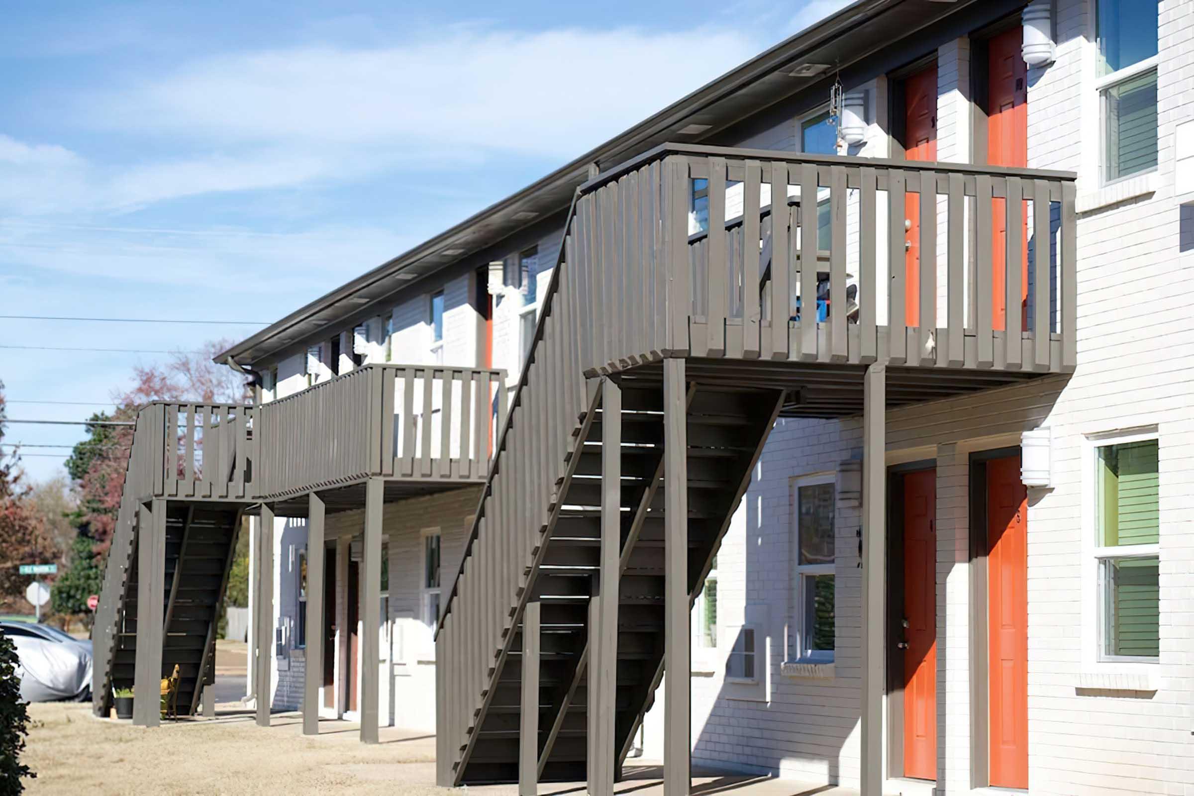 A view of a two-story apartment building featuring exterior stairs leading to upper units. The building has a modern design with white brick walls and brightly colored doors in shades of orange. A grassy area is visible in front, with a clear blue sky overhead.