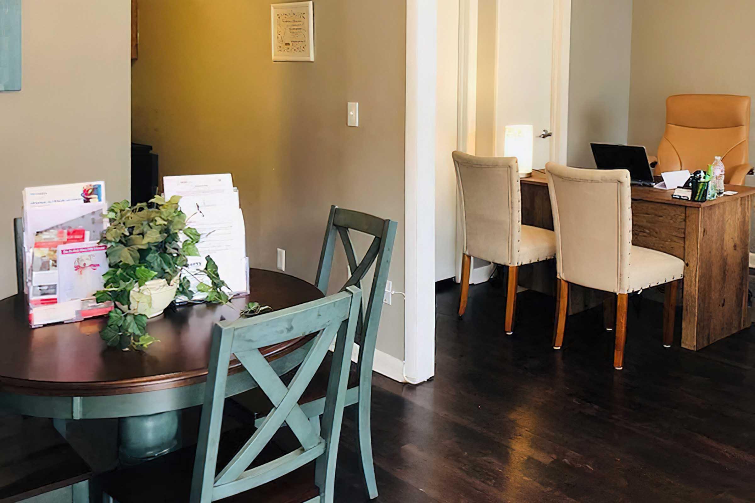 A cozy office space featuring a round table with chairs and a decorative plant in the foreground, and a desk with a computer and an orange chair in the background. The walls are painted in a warm tone, creating a welcoming atmosphere. Papers and brochures are arranged neatly on the table.